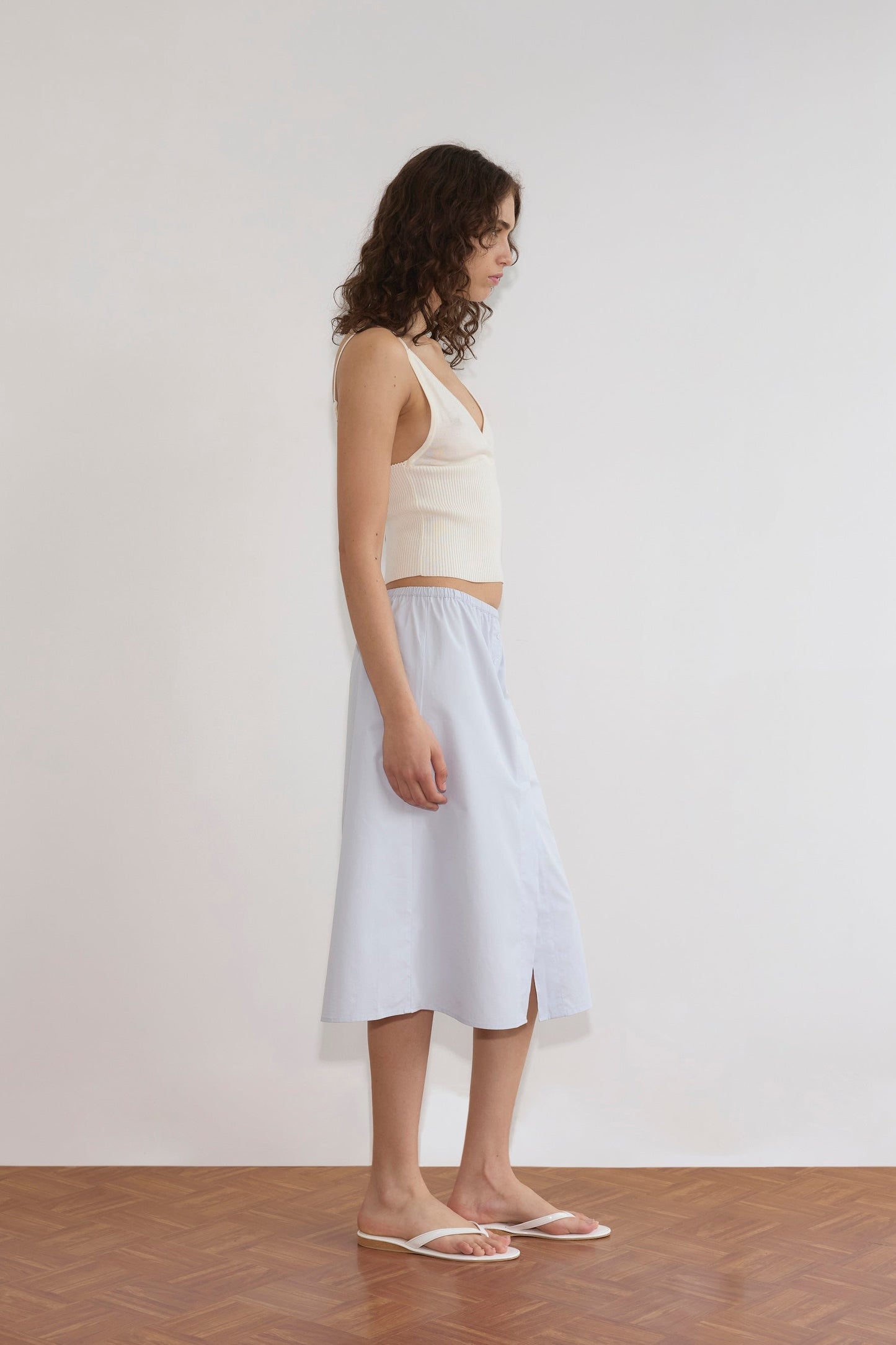 A woman with curly brown hair stands in profile against a plain white wall, wearing a Cross Panel Knit Top - Off White, a light blue midi skirt with a front slit, and white sandals on a wooden floor.