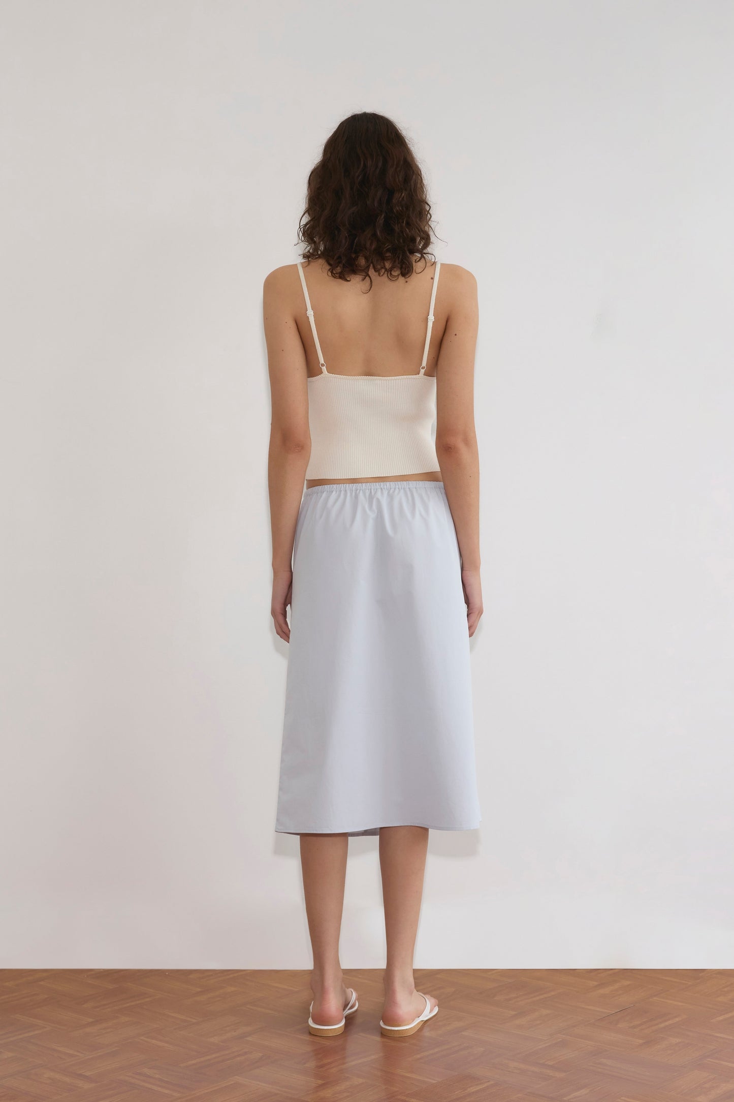 A woman with curly hair faces a white wall, wearing an off-white Cross Panel Knit Top, a light blue midi skirt, and white sandals on a wooden floor.