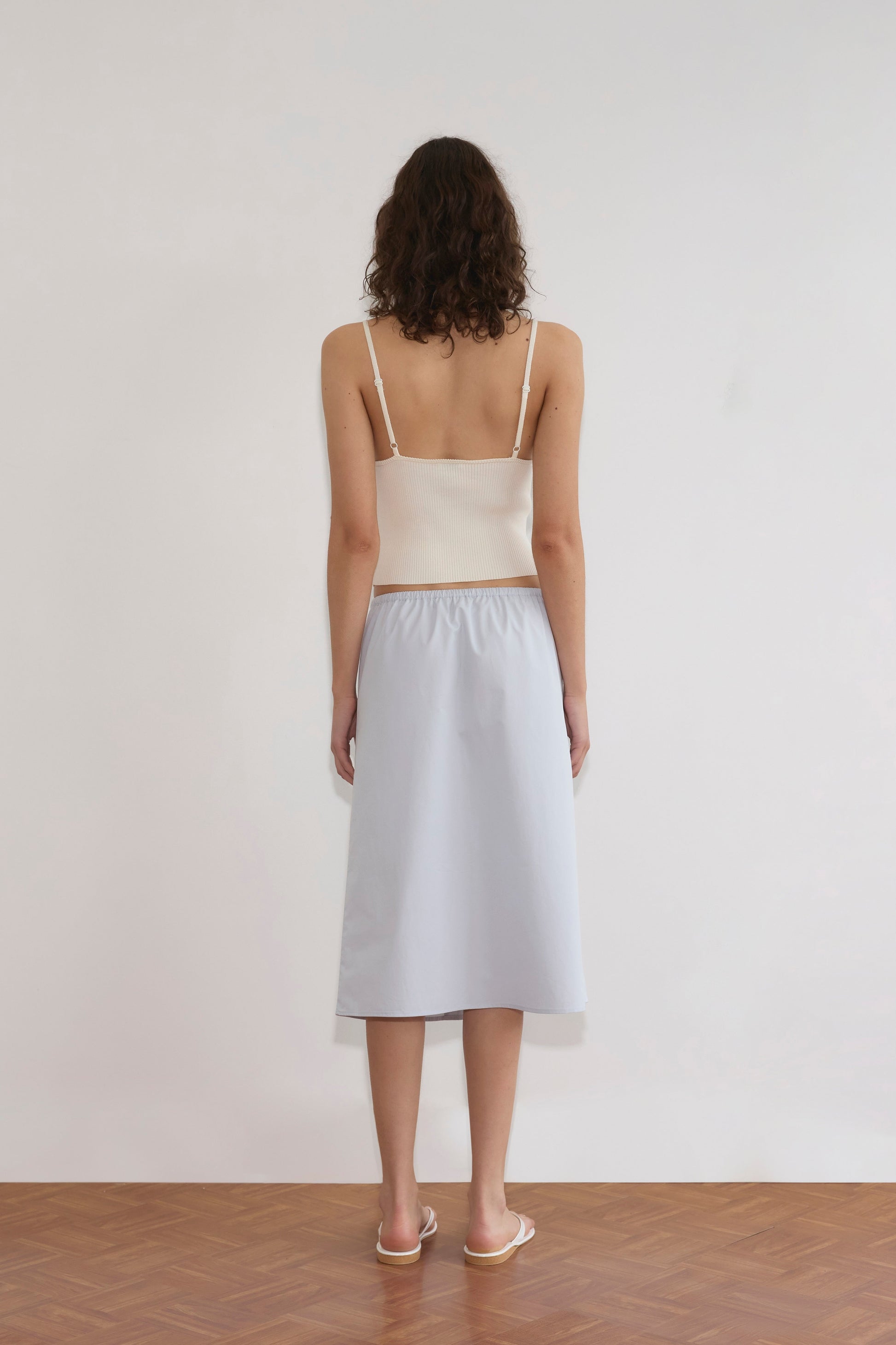 A woman with curly hair faces a white wall, wearing an off-white Cross Panel Knit Top, a light blue midi skirt, and white sandals on a wooden floor.