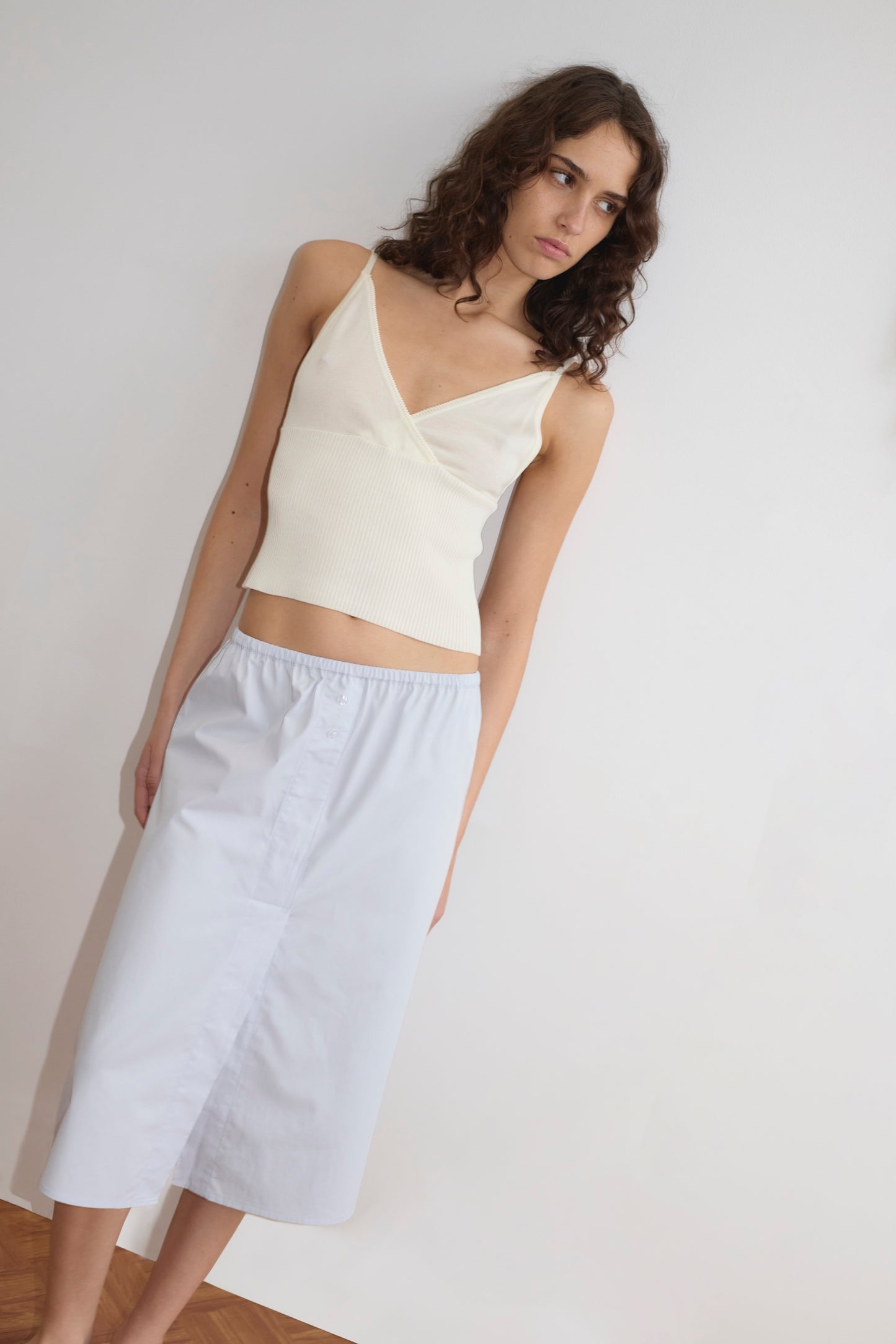 A woman with wavy brown hair wears a Deiji Studios Cross Panel Knit Top in Off White and a light blue midi skirt, standing against a plain white wall and looking slightly to the side.