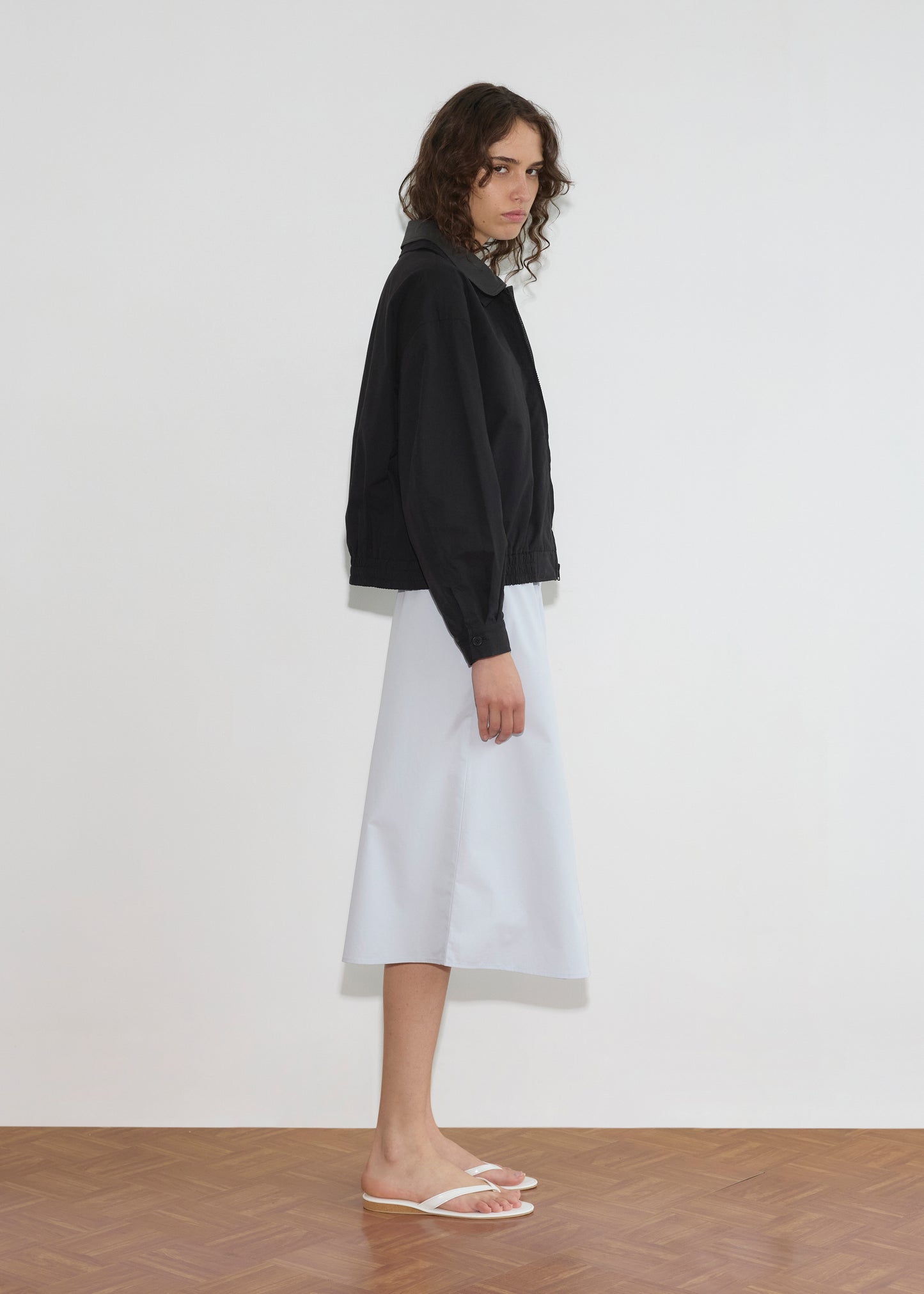 A person with wavy, shoulder-length hair stands sideways against a white wall, wearing the Collared Zip Jacket - Black Contrast over a white midi skirt and white flip-flops on a wooden floor.