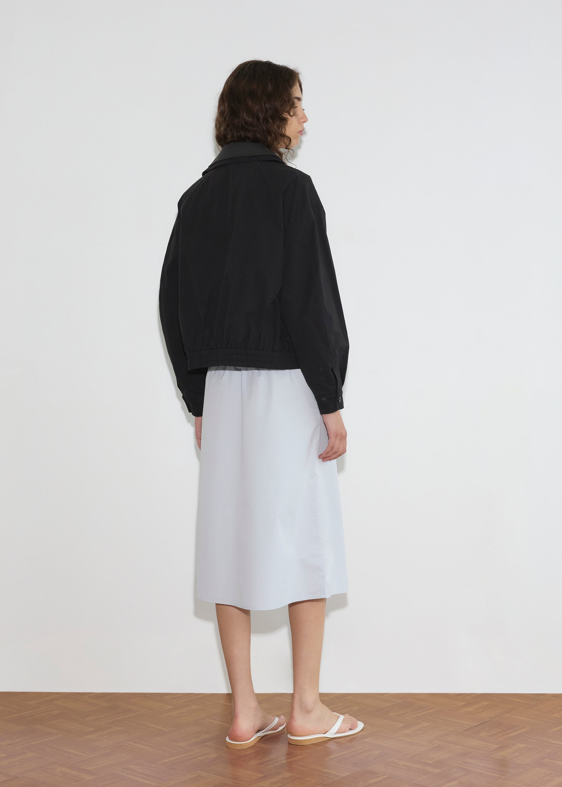 A person with curly hair faces a white wall, wearing the Collared Zip Jacket - Black Contrast, a light blue knee-length skirt, and white sandals on a parquet floor.