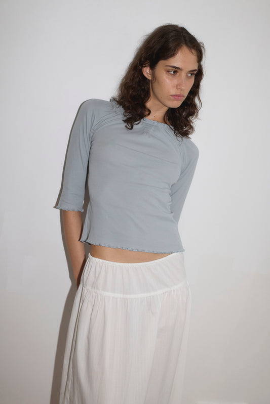 A young woman with wavy brown hair wears the Cotton Flounce 3/4 Sleeve - Baby Blue top and a white skirt, standing against a plain white wall and looking slightly to the side with a neutral expression.