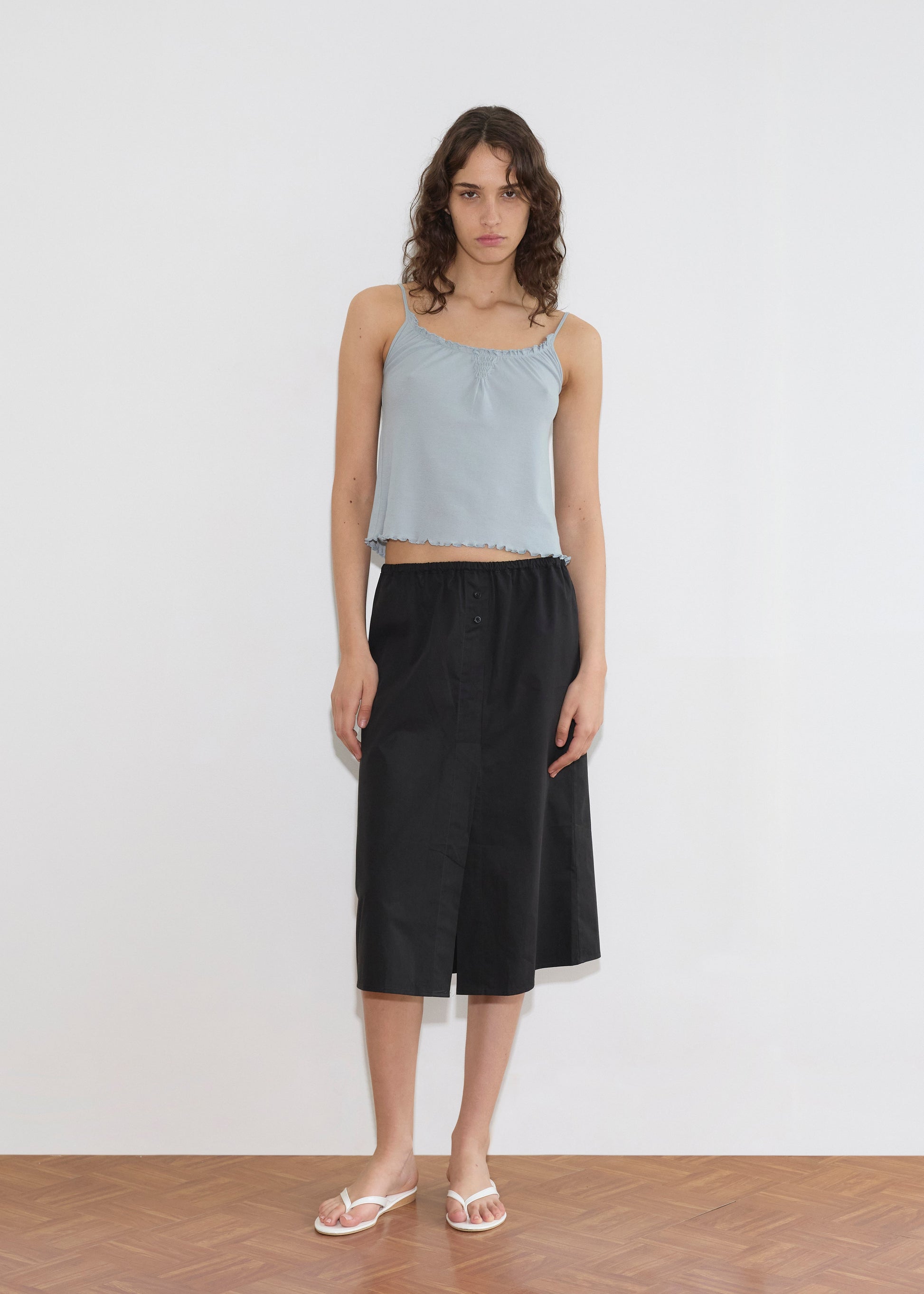 A model wearing a light blue cropped camisole with ruffled trim and the Overlap Skirt—a black midi skirt with an elasticated waist and front slit—paired with white flip-flops, stands against a white wall.