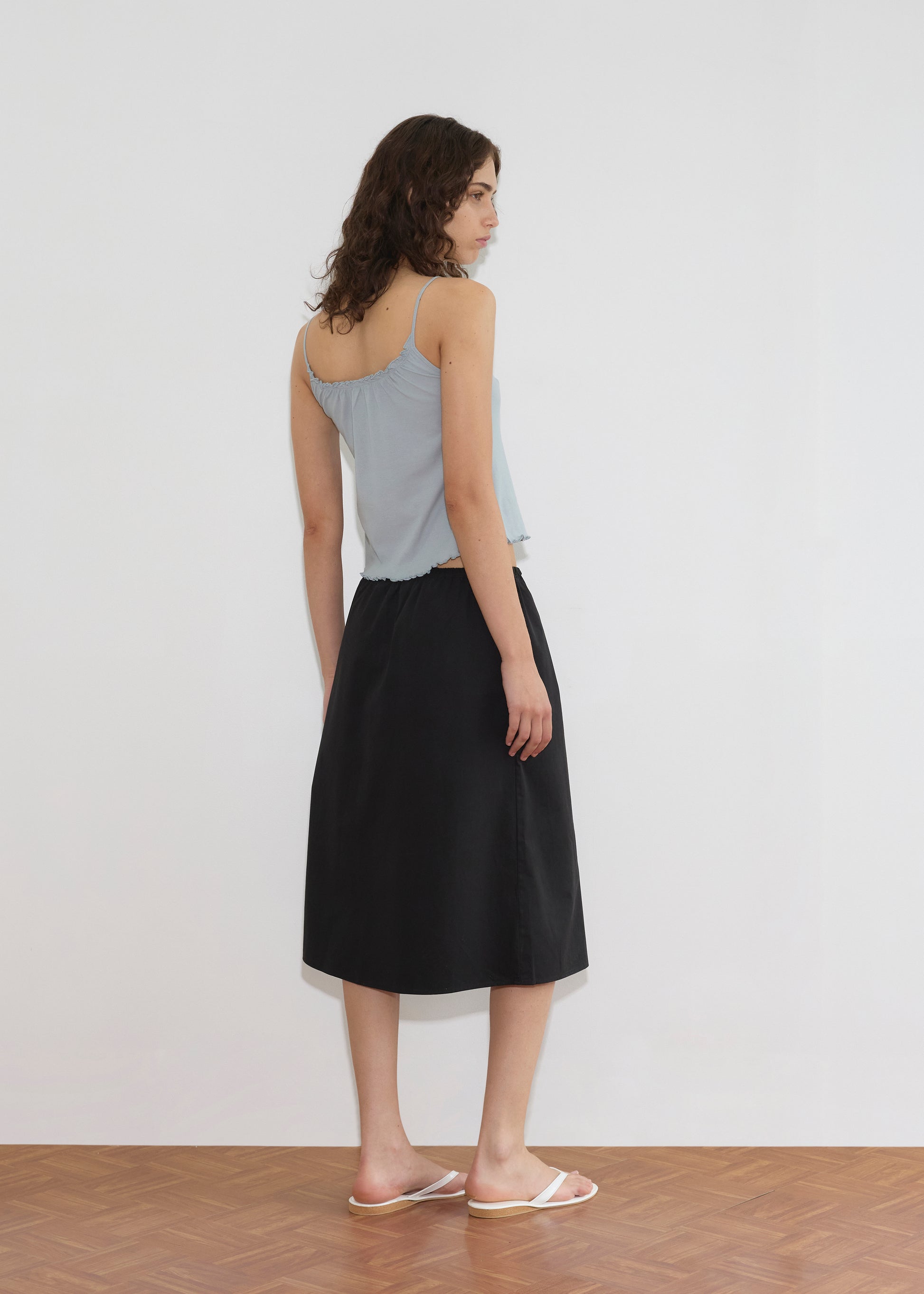 A woman with wavy brown hair stands facing away, wearing the Cotton Flounce Tank - Baby Blue by Deiji Studios, a black midi skirt, and white sandals on a wooden floor against a plain white wall.