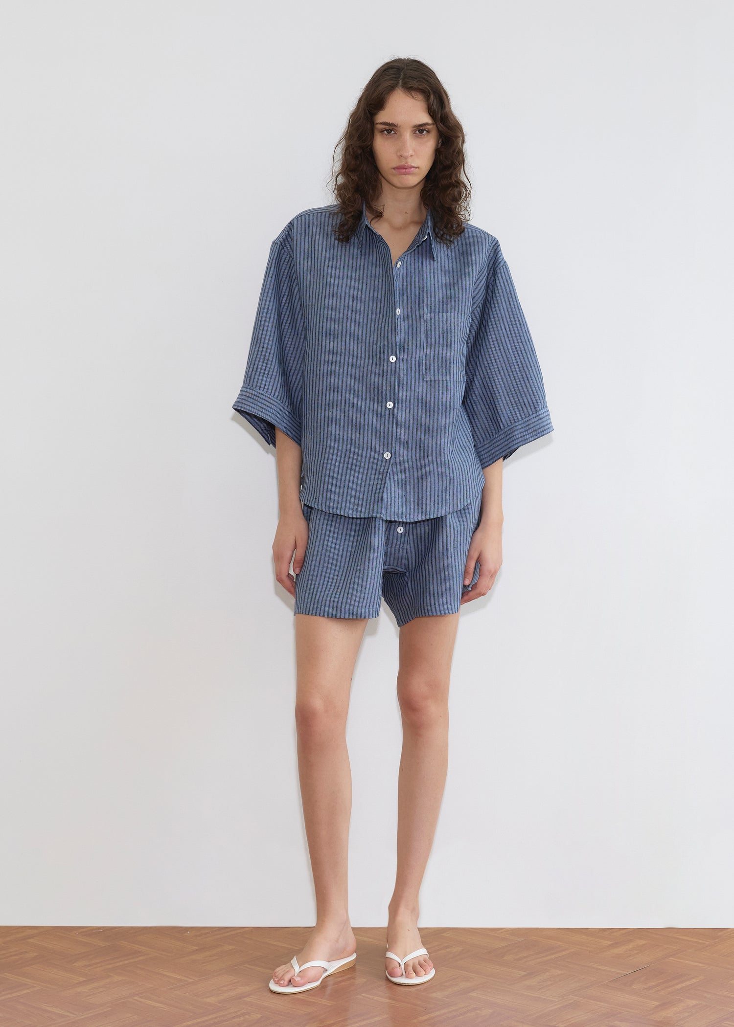 A person with long wavy hair wears the 03 Set - Azure Stripe by Deiji Studios—loose linen shirt and matching boxer shorts—standing barefoot in white flip-flops on a wooden floor against a plain white wall.