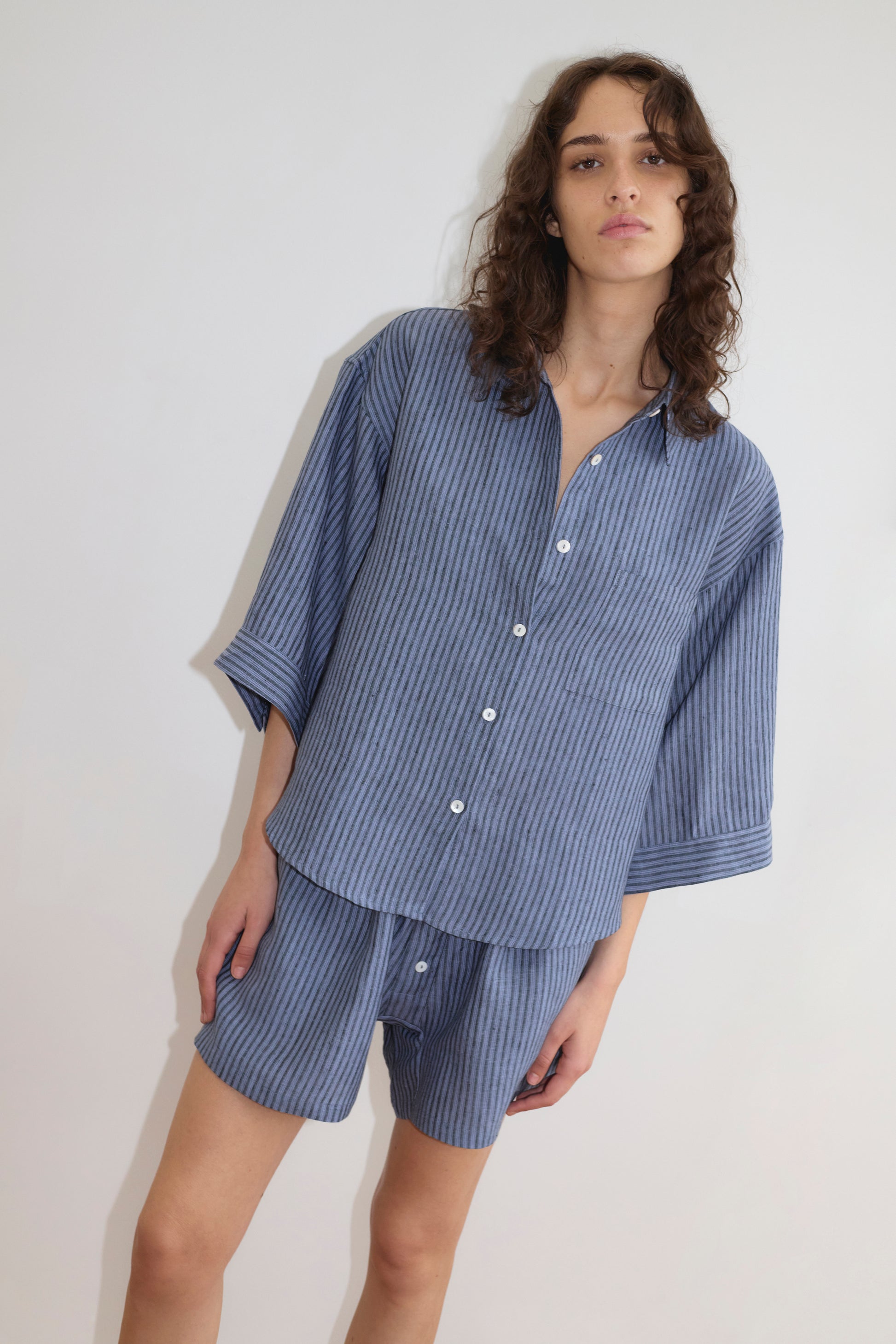 A person with wavy brown hair wears the 03 Set - Azure Stripe by Deiji Studios, featuring a loose-fitting blue and white pinstriped linen shirt and matching boxer shorts, standing against a plain white background.