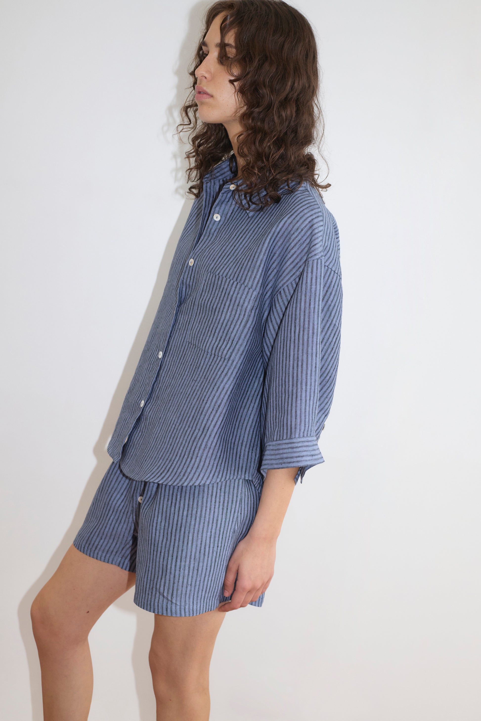 A person with wavy brown hair wears the Deiji Studios 03 Set - Azure Stripe—blue and white vertically striped linen shirt and matching boxer shorts—standing against a plain white background.