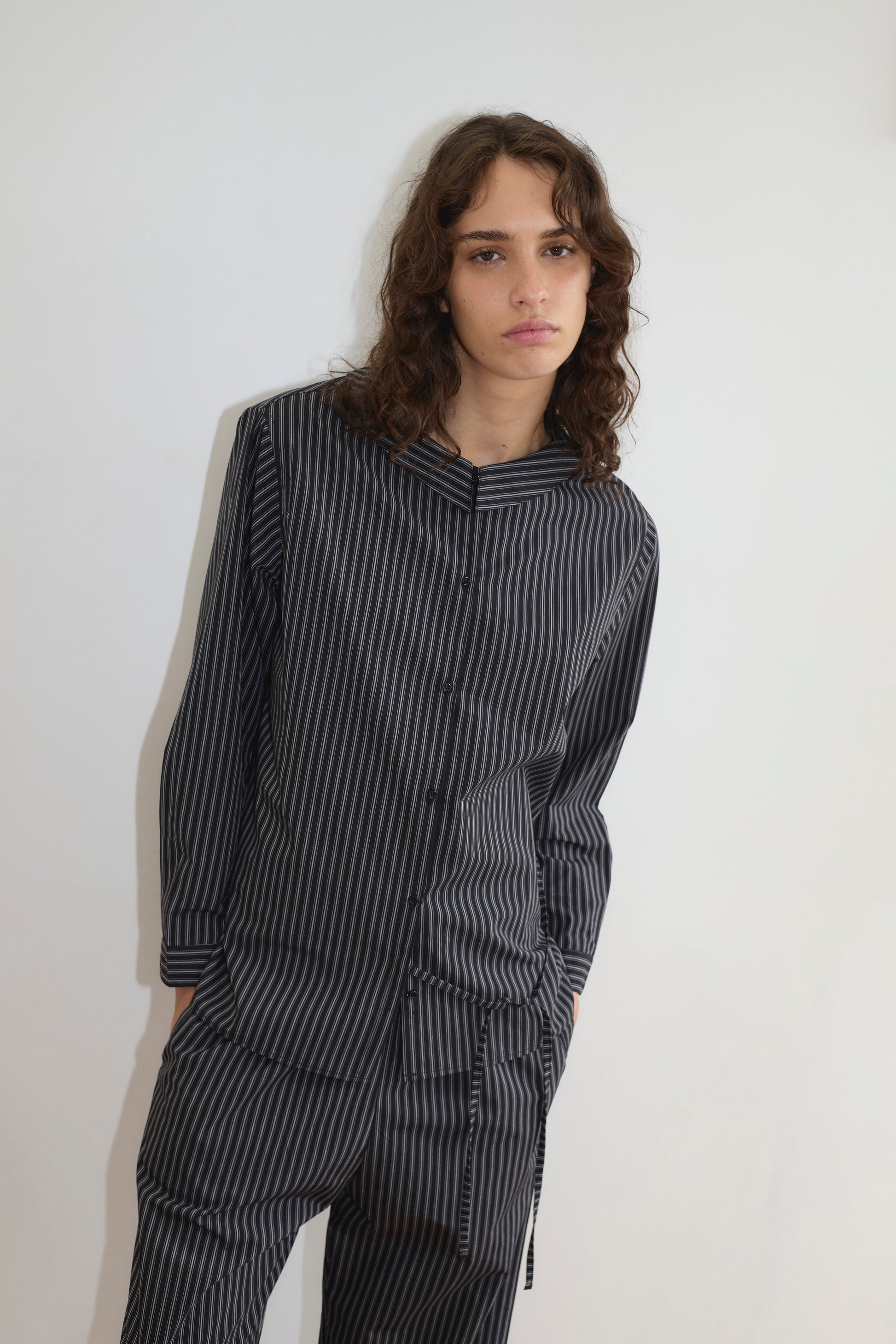 A person with long, wavy brown hair wears the Left Set - Mono Stripe by Deiji Studios—a relaxed, black and gray vertically striped pajama-style suit—and stands calmly against a plain white wall, looking at the camera.