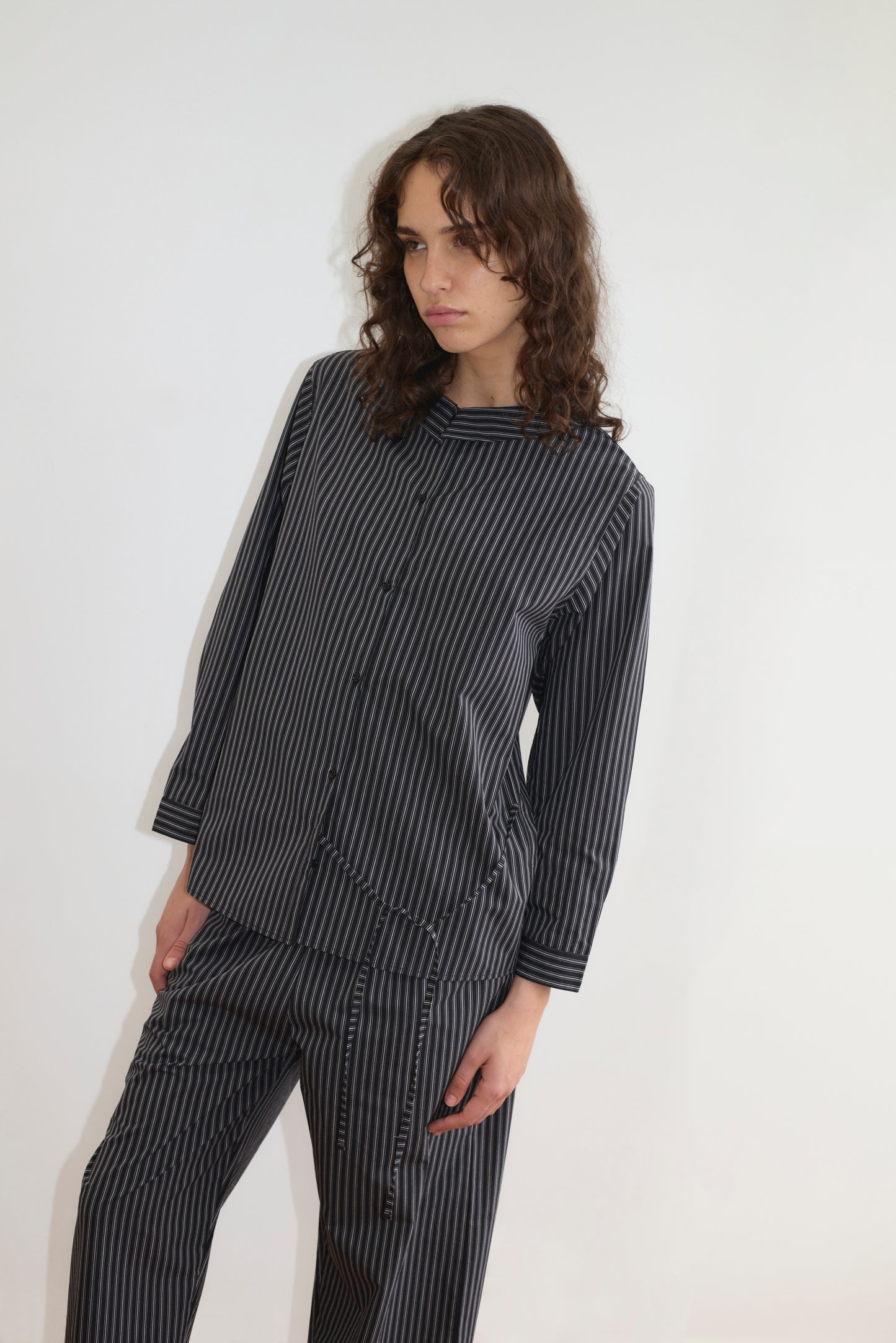 A person with wavy brown hair wears the Deiji Studios Left Set - Mono Stripe: a matching black and gray vertical striped lightweight shirt and pants, standing against a plain white background, looking slightly to the side with a neutral expression.