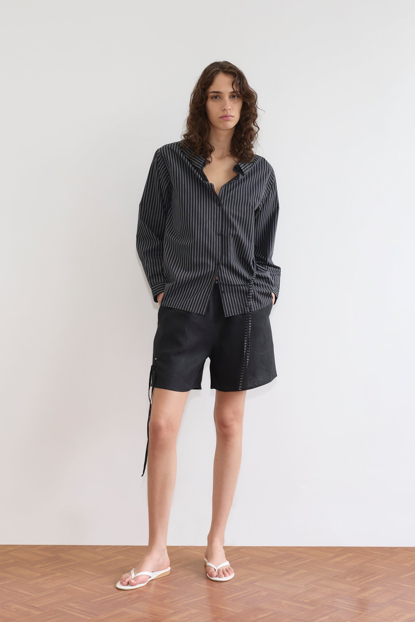A person stands against a plain white wall in a relaxed pose, wearing the Deiji Studios Left Shirt - Mono Stripe, loose black shorts with visible stitching, and white flip-flops. Their hands are in their pockets and they have shoulder-length wavy hair.