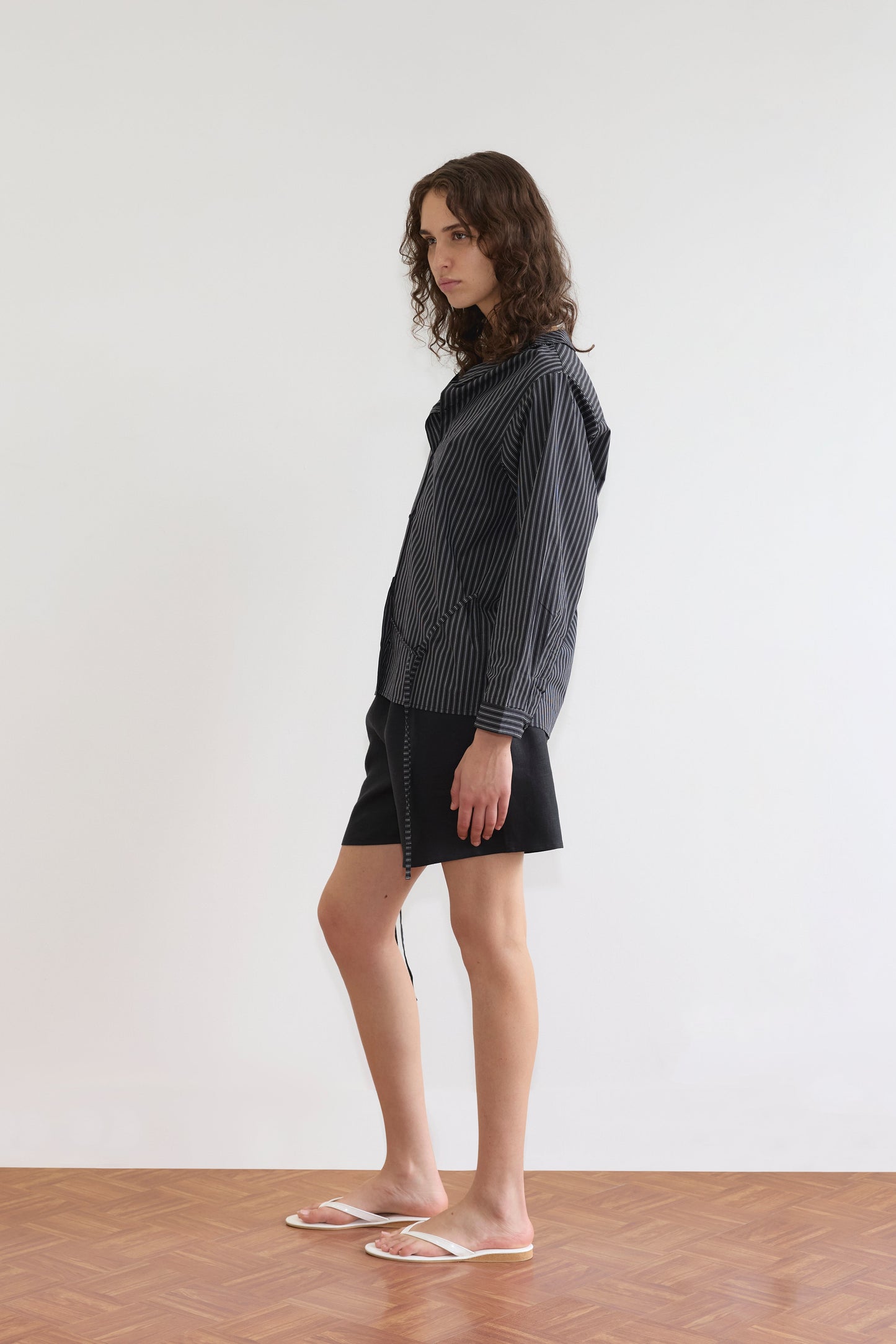 A person with wavy brown hair stands sideways on a wooden floor by a white wall, wearing the Deiji Studios Left Shirt - Mono Stripe (long-sleeved, black-and-white striped), black shorts, and clear sandals.