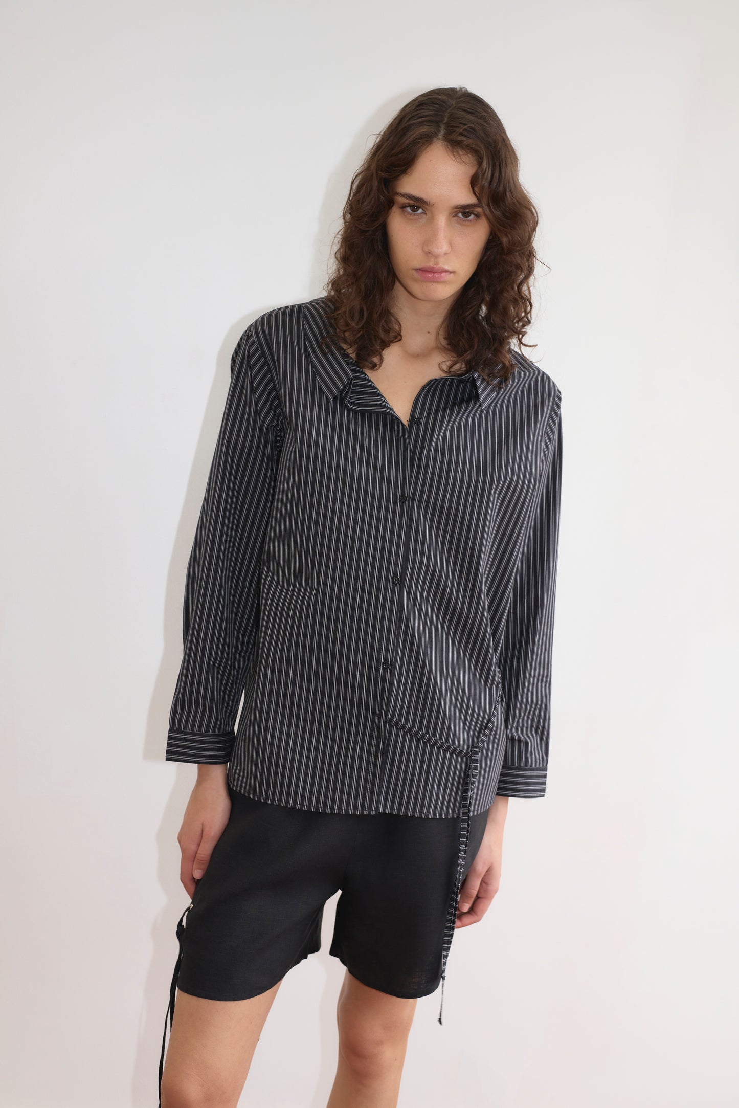 A person with long, wavy brown hair wears the Deiji Studios Left Shirt - Mono Stripe, a lightweight vertically striped button-up with a relaxed fit, paired with black shorts and standing against a white background with a serious expression.