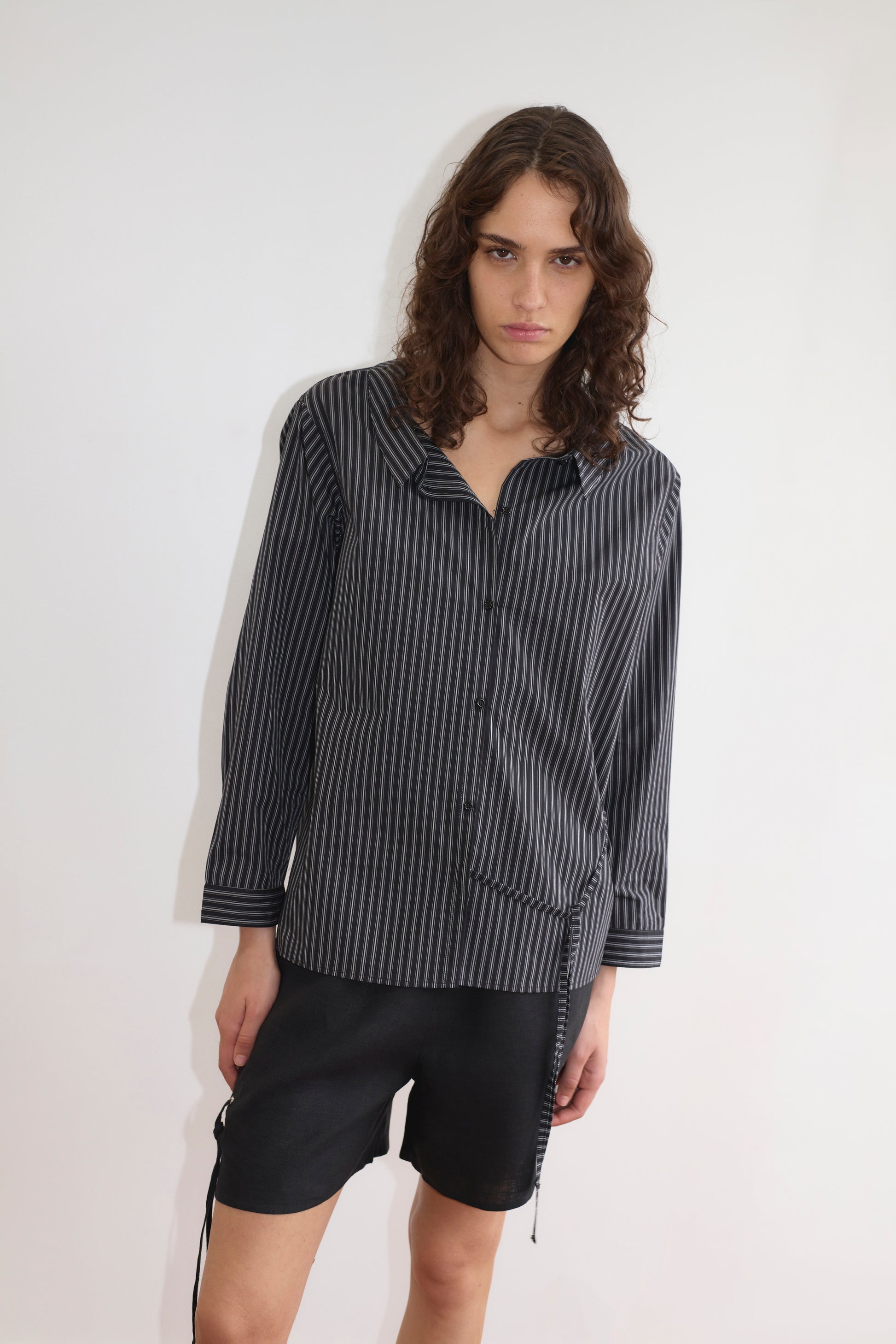 A person with long, wavy brown hair wears the Deiji Studios Left Shirt - Mono Stripe, a lightweight vertically striped button-up with a relaxed fit, paired with black shorts and standing against a white background with a serious expression.