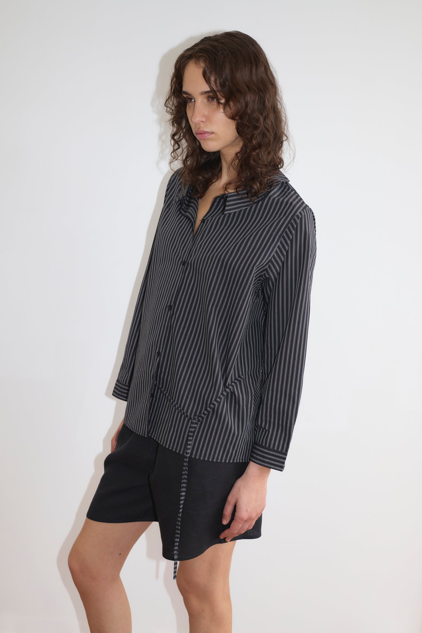 A person with long curly hair stands against a white background, wearing the Left Shirt - Mono Stripe by Deiji Studios and black shorts, looking slightly to the side with a neutral expression and relaxed silhouette.