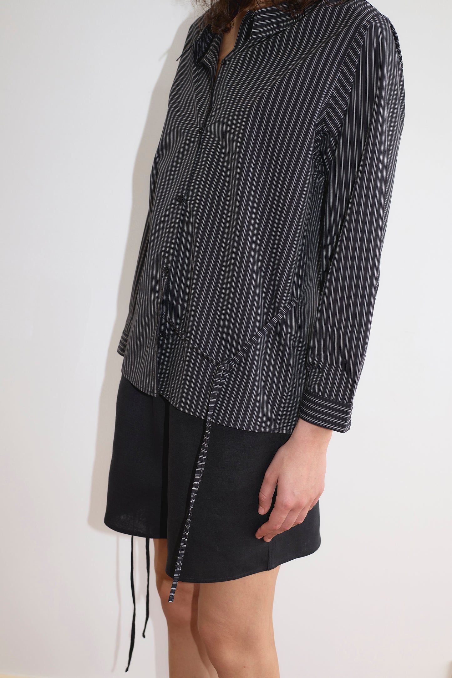 A person wears the Deiji Studios Left Shirt - Mono Stripe, a black pinstriped long-sleeve blouse with a relaxed fit and waist tie, paired with a black skirt against a plain white background. The person's face is not visible.