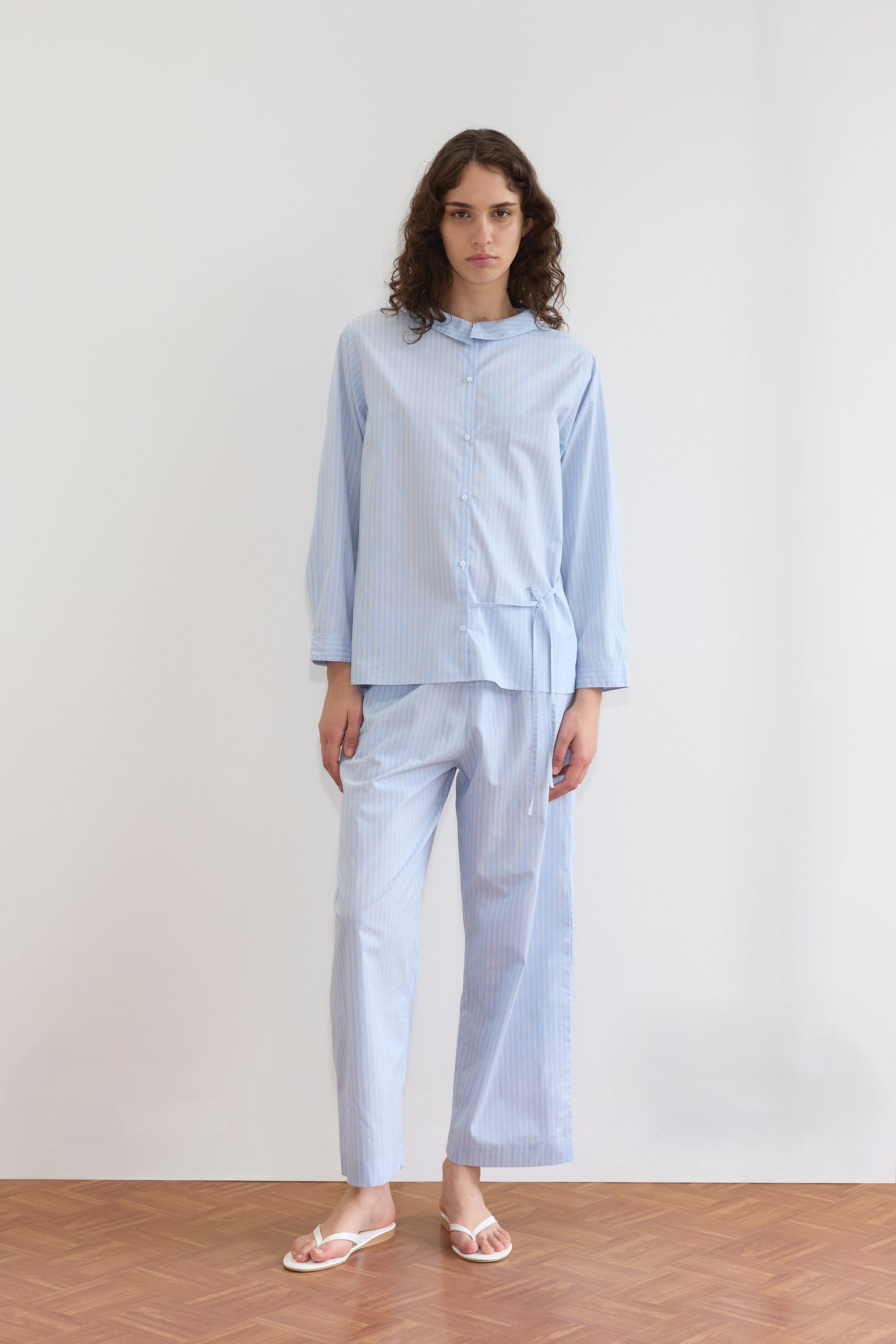 A person with long curly hair stands indoors in a relaxed pose, wearing the Left Set - Lake Blue Stripe, featuring lightweight, elastic waist pants and white flip-flops, against a plain white wall and wooden floor.