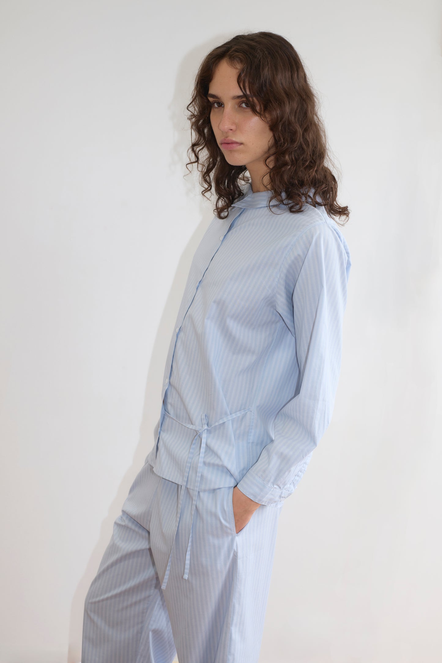 A person with long, wavy brown hair stands against a white background wearing the Left Set - Lake Blue Stripe, a light blue pinstripe matching set with a lightweight shirt and belted elastic waist pants, looking neutrally at the camera.