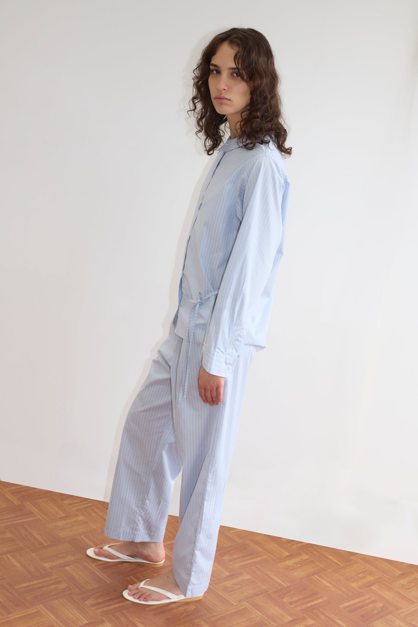 A person with curly brown hair stands sideways on a wooden floor, looking toward the camera while wearing the Left Set - Lake Blue Stripe: a loose-fitting lightweight shirt and matching elastic waist pants, paired with white sandals.