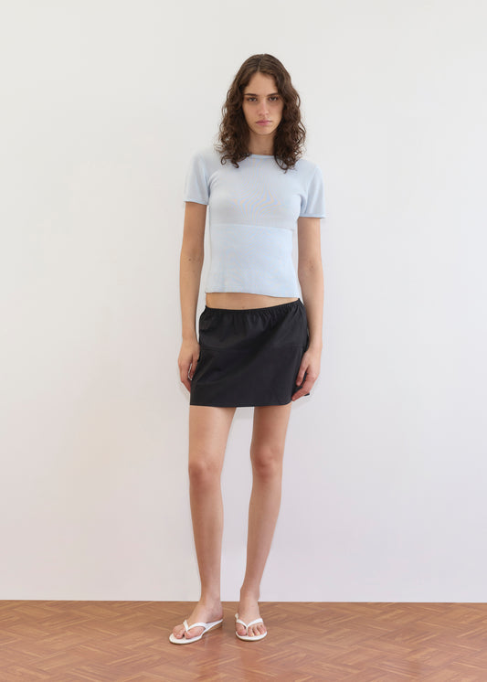 A person with wavy brown hair stands against a plain white wall, wearing a light blue short-sleeve top, Deiji Studios' Pocket Skirt - Black (mini-length), and white sandals on a light brown parquet floor.