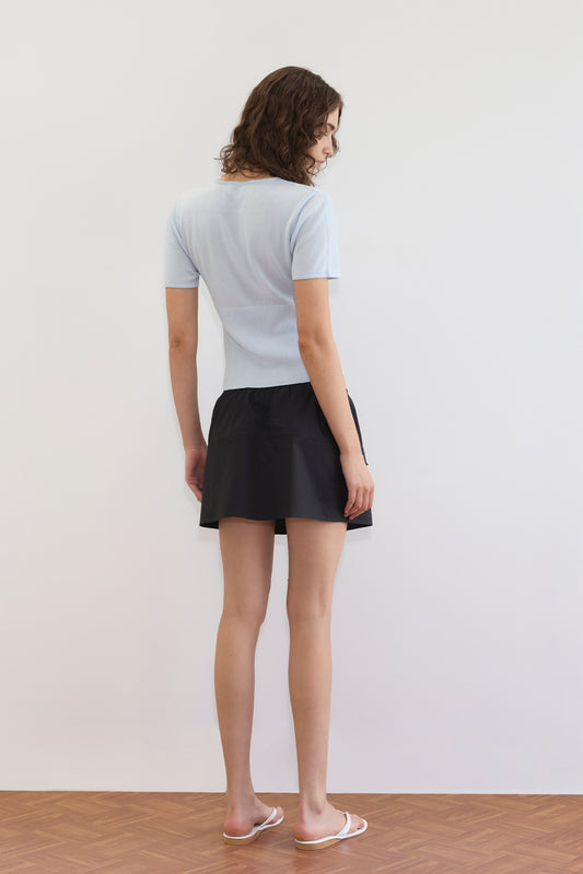 A woman with curly hair faces away from the camera, wearing a light blue short-sleeve top, the Pocket Skirt - Black by Deiji Studios, and white sandals, against a plain white wall and wood-patterned floor.