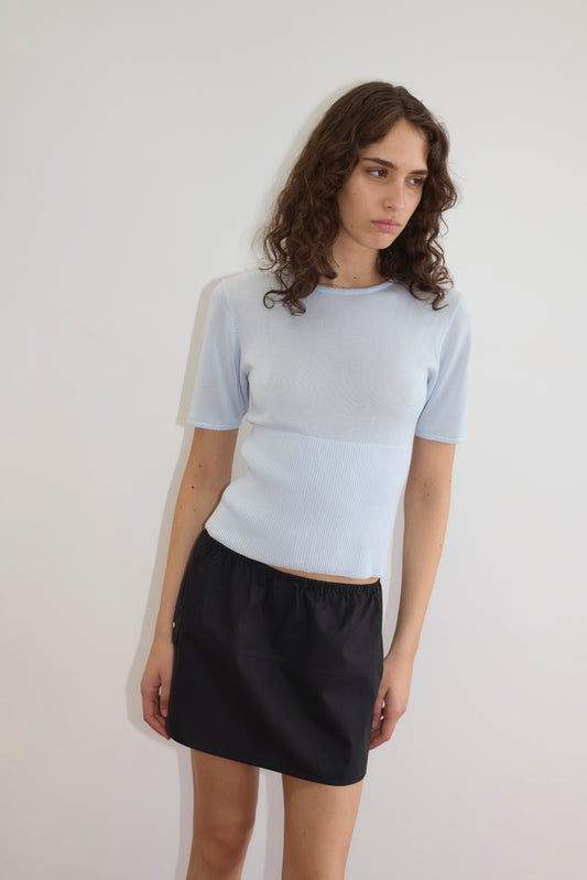 A person with wavy brown hair in a light blue short-sleeve top and the Deiji Studios Pocket Skirt - Black (mini length) stands against a plain white wall, gazing slightly down and to the side.