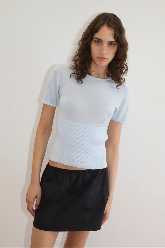A woman with wavy brown hair wears the Trim Knit Tee - Ice Blue and a black skirt, standing against a white background. She faces the camera with a neutral expression.