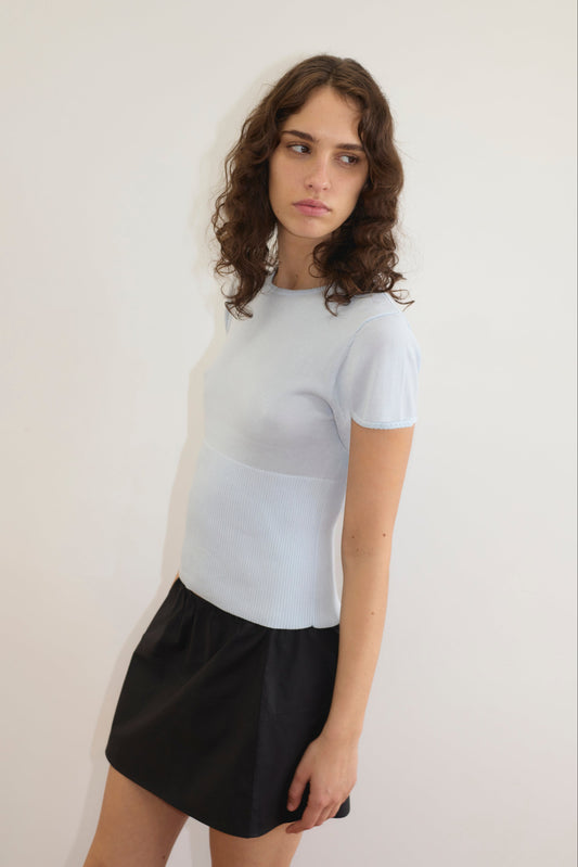 A woman with wavy brown hair wears the Trim Knit Tee - Ice Blue and a black skirt, standing against a plain white wall and looking to the side with a neutral expression.
