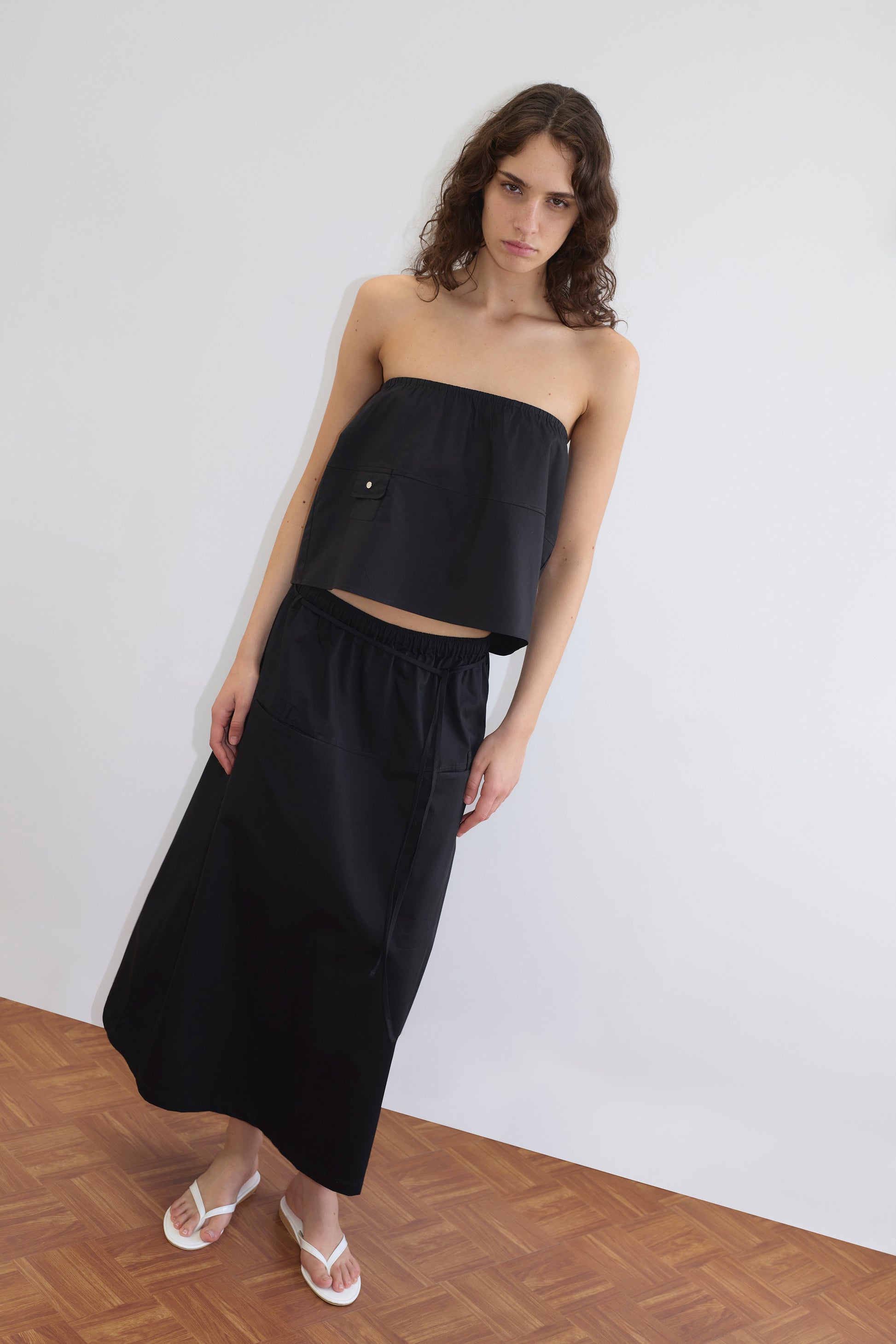 Model wears a Deiji Studios black strapless crop top with a chest pocket, paired with the Loop Pocket Skirt - Black featuring an elastic waistband, and white thong sandals, standing against a white wall.