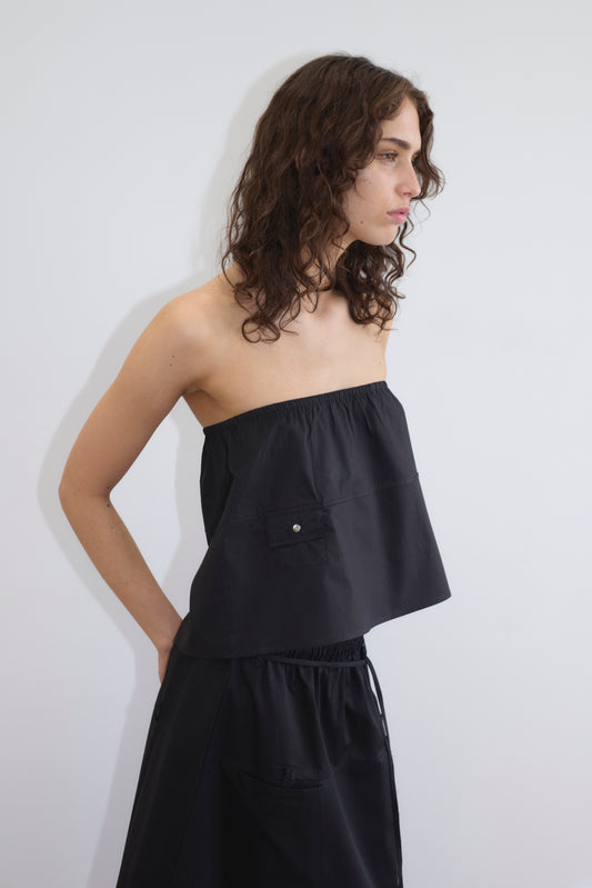 A person with wavy brown hair wears a black, strapless, loose top and the Pocket Skirt - Black by Deiji Studios, standing against a plain white background and looking to the right.