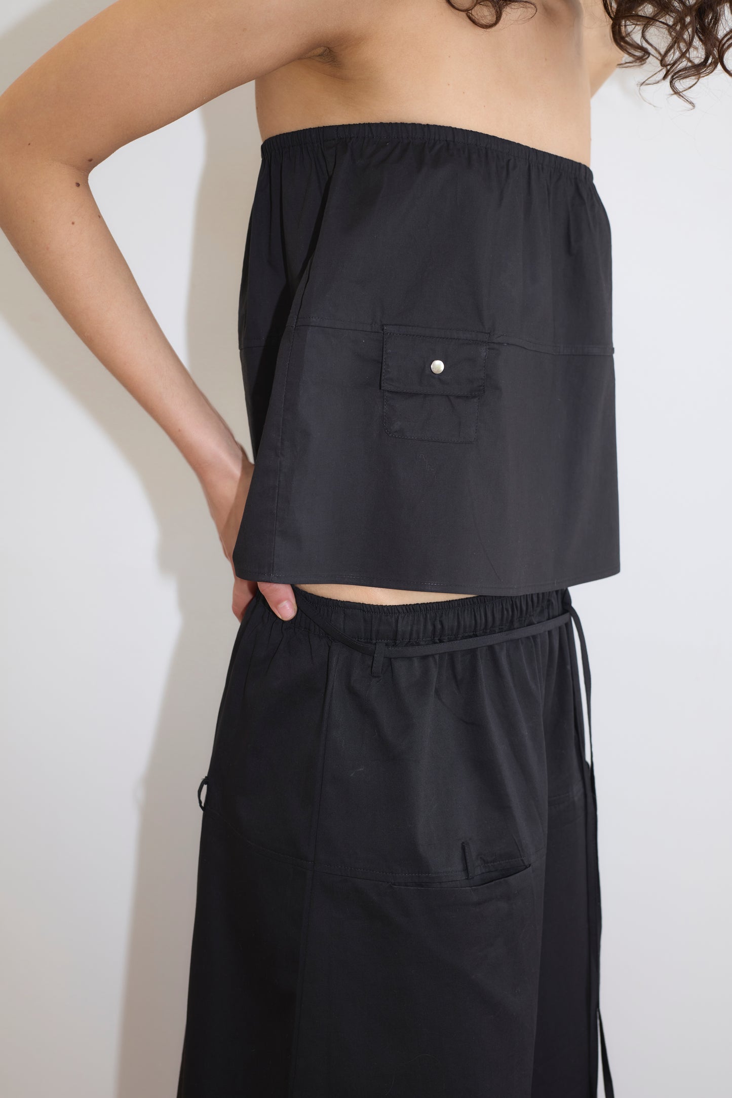 A person wears the Deiji Studios Pocket Skirt in black, featuring a loose fit and pocket, paired with matching black pants with an elastic waistband. They stand against a white background with arms bent at the elbows.