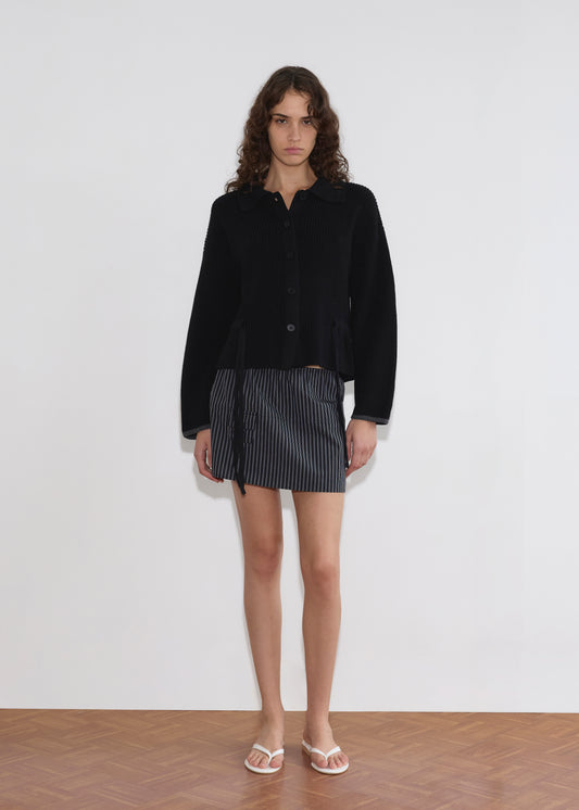 A person with long curly hair stands indoors against a plain white wall, wearing the black Tie Side Cardigan and a short black-and-white pinstripe skirt, plus white sandals on a wooden floor.