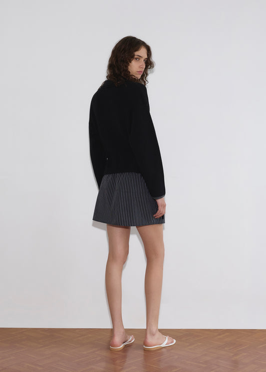 A person with long curly hair stands facing away, looking over their shoulder. They wear the Tie Side Cardigan - Black by Deiji Studios, a short pinstriped skirt, and white flip-flops on a wooden floor against a plain white wall.