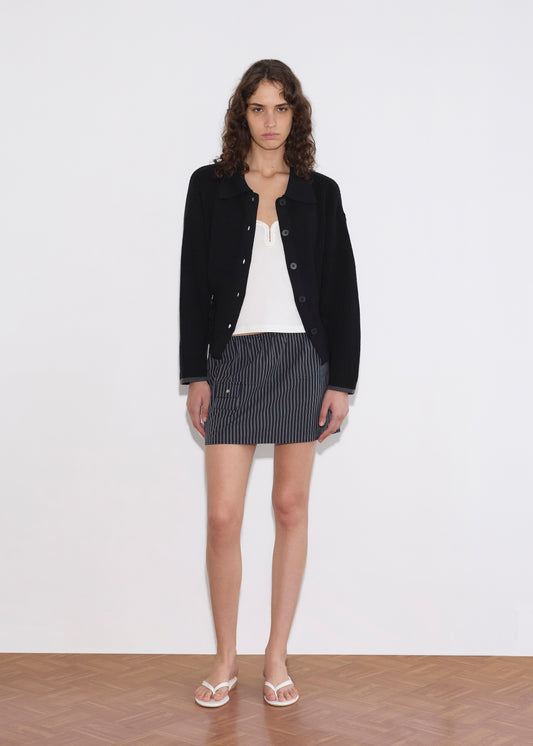 Wearing Deiji Studios' Tie Side Cardigan in black over a white top, a gray pinstripe mini skirt, and white flip-flops, a person with curly hair stands against a white wall on wooden flooring for an effortless look.