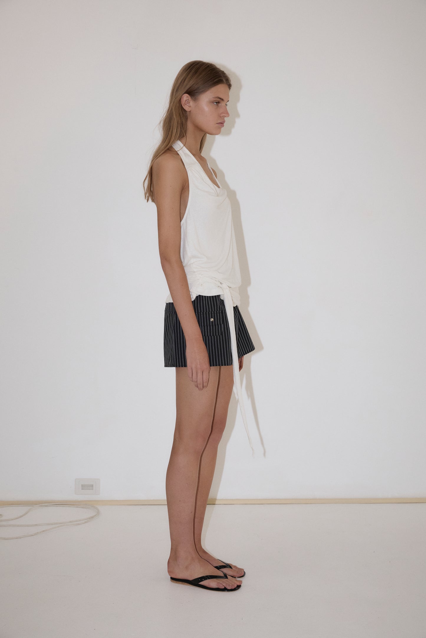 A woman stands in profile against a plain white wall, wearing Deiji Studios’ Drape Halter - White, black pinstriped shorts, and black flip-flops. Her long hair is loose, and a power outlet is visible on the wall behind her.