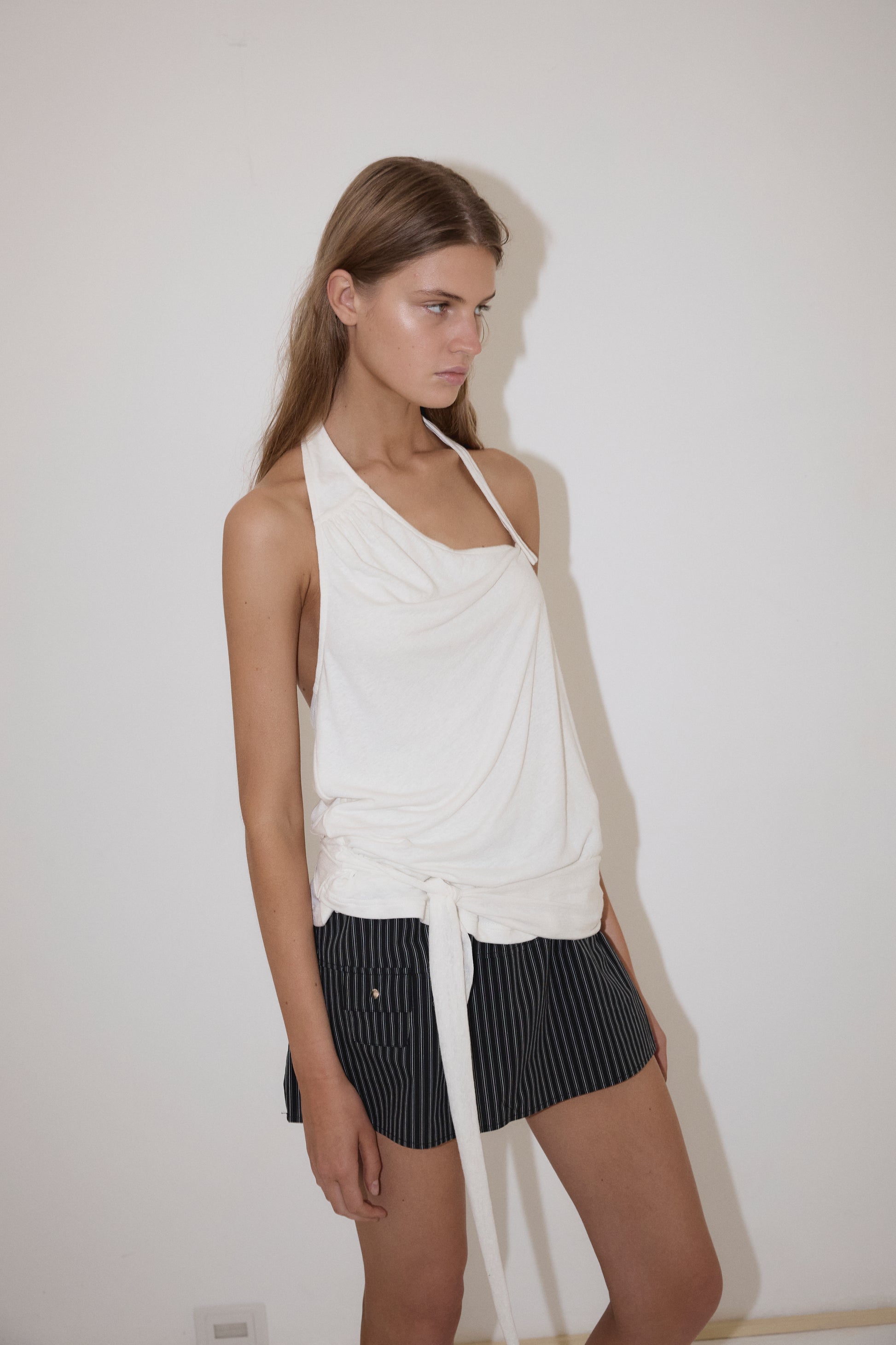 A woman with straight brown hair and a neutral expression stands against a white wall, wearing the Drape Halter - White top by Deiji Studios paired with dark pinstriped shorts.