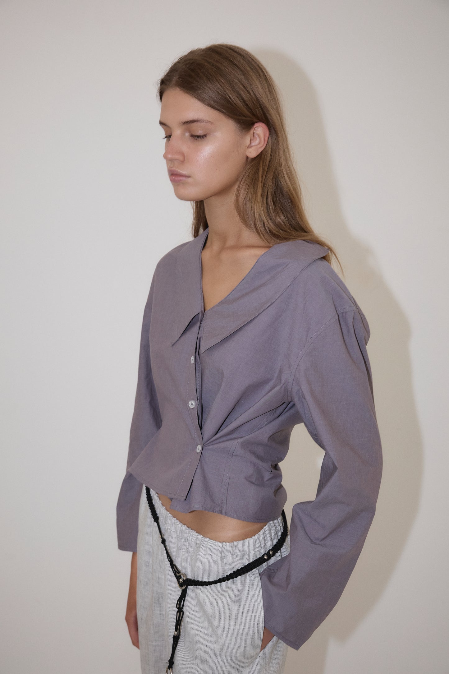 A young woman with long brown hair stands before a white wall, wearing a Deiji Studios Waist Button Shirt in Currant and light gray high-waisted pants, looking to the side with a neutral expression.