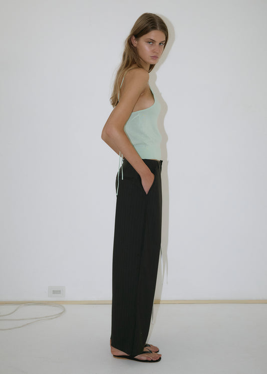 A woman with long hair stands in profile against a plain white wall, wearing a light green sleeveless top, the Bind Pant in Charred Black Stripe by Deiji Studios, and black sandals, with her hands in her pockets.