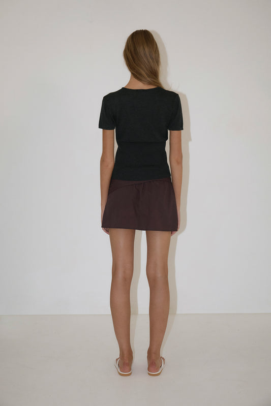 A woman with long hair faces away from the camera, wearing a Trim Knit Tee - Dark Grey by Deiji Studios, a dark brown mini skirt, and white flip-flops against a plain white wall and floor.