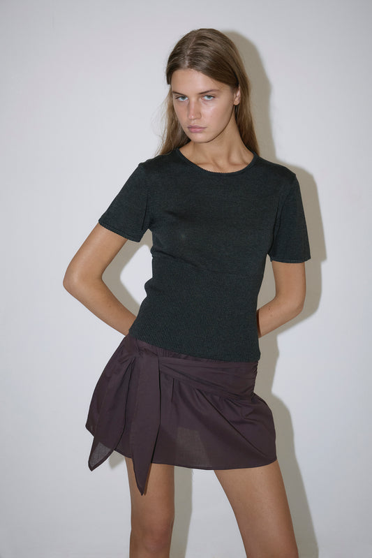 A woman with long brown hair stands against a plain white wall wearing the Trim Knit Tee in dark grey and a dark mini skirt with a tied detail. She has a serious expression and one hand on her hip.