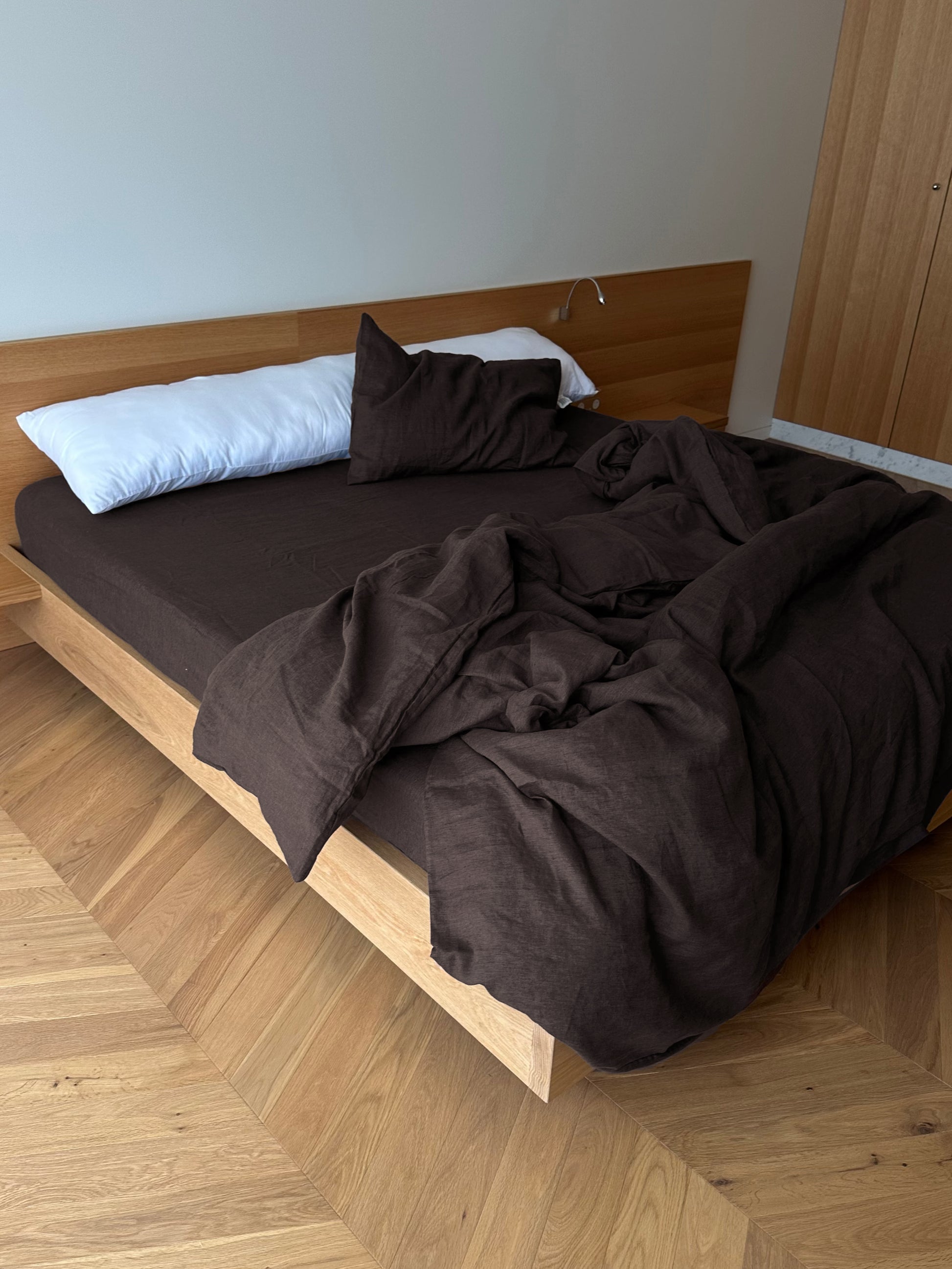 A modern wooden bed is dressed in the Duvet cover - Bark, a dark brown blanket, matching pillow, and a long white hypo-allergenic pillow. Cozy linen and minimal decor on light wood floors capture the signature Deiji Studios style.