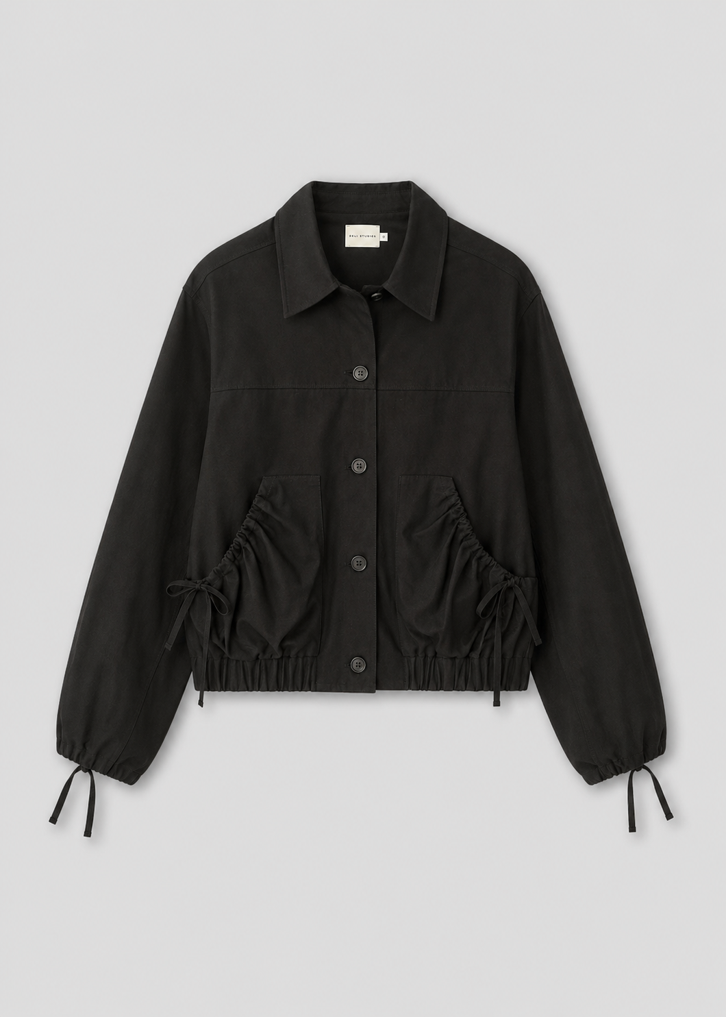 The Button Up Jacket - Black features a collar, two front pockets with gathered drawstring details, and elastic cuffs with tie accents on the sleeves. Inspired by Deiji Studios, this cropped jacket has a relaxed, casual style.