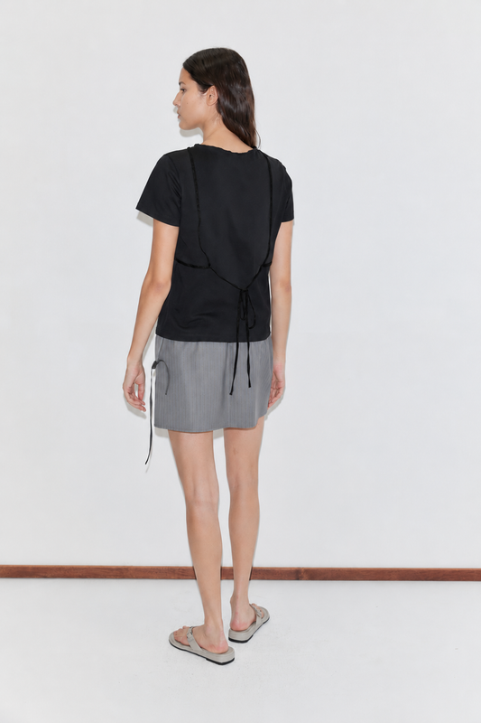 A woman faces away, wearing the Tape Tee - Black by Deiji Studios, a gray mini skirt with a white tie detail, and flip-flops. Her dark hair is down against a plain white background.