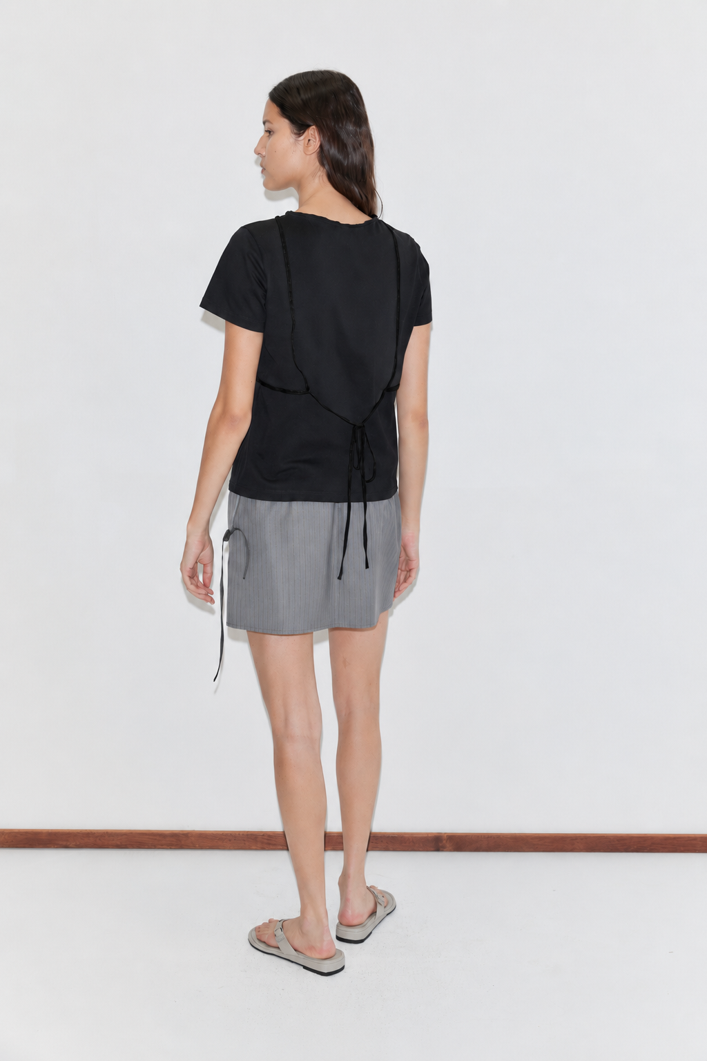 A woman stands with her back to the camera, wearing a black short-sleeve top with back ties, the Wool Tie Mini Skirt - Grey Stripe, and gray flip-flops, all set against a plain white background.