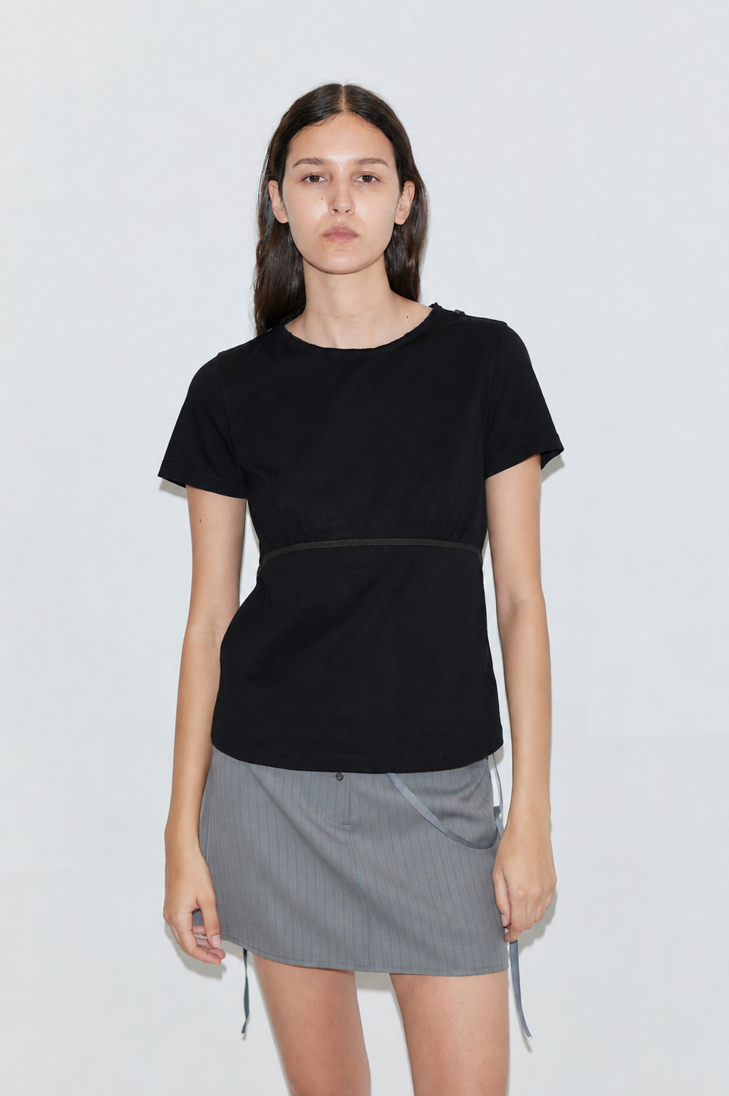 A young woman with long dark hair stands against a plain backdrop, wearing the Tape Tee - Black and a gray pinstriped mini skirt. She faces the camera with a neutral expression.