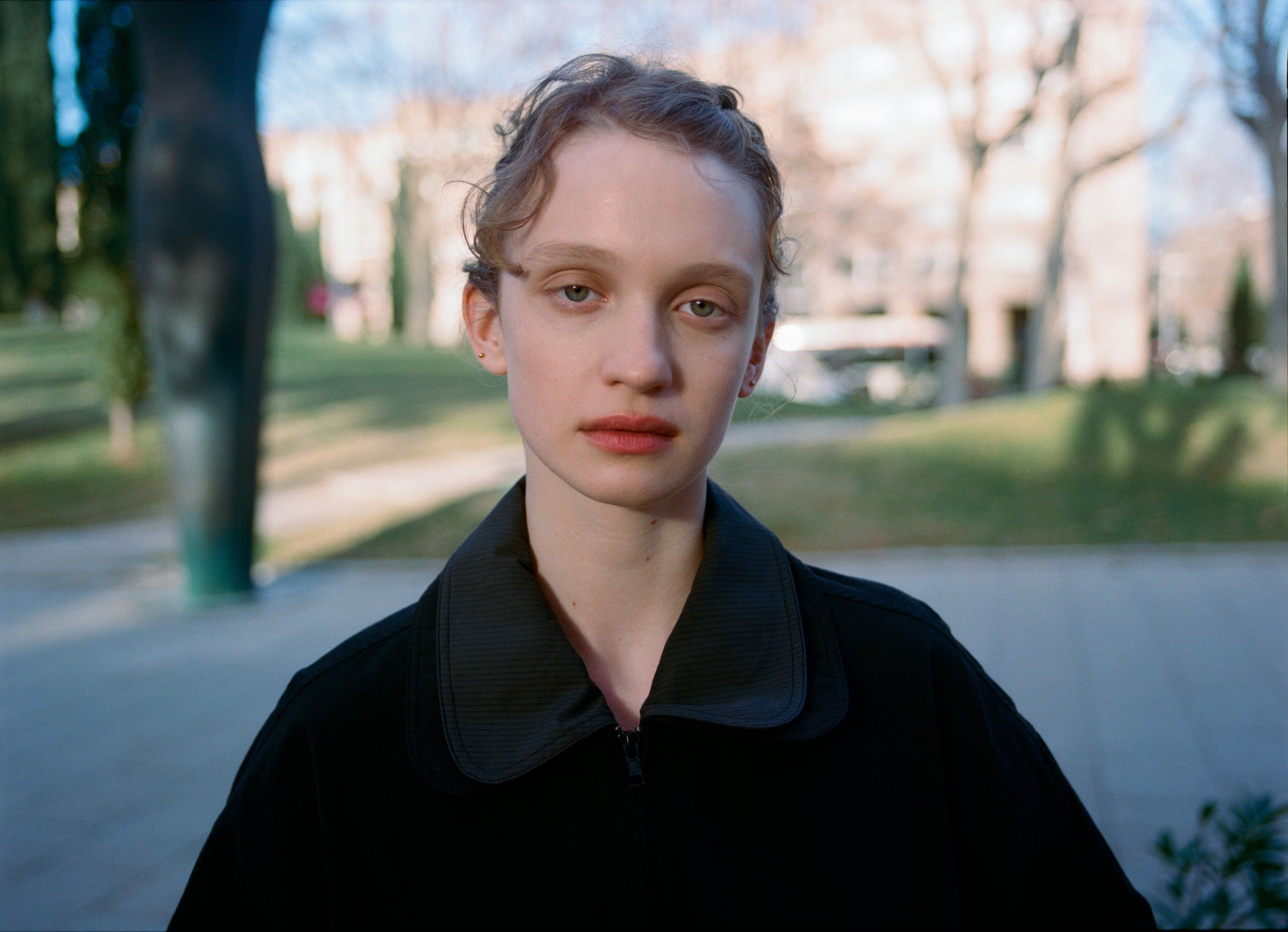 A young person with short, curly hair and a neutral expression stands outdoors in a park, wearing the Collared Zip Jacket - Black Contrast. Leafless trees and blurred buildings are visible in the sunny background.