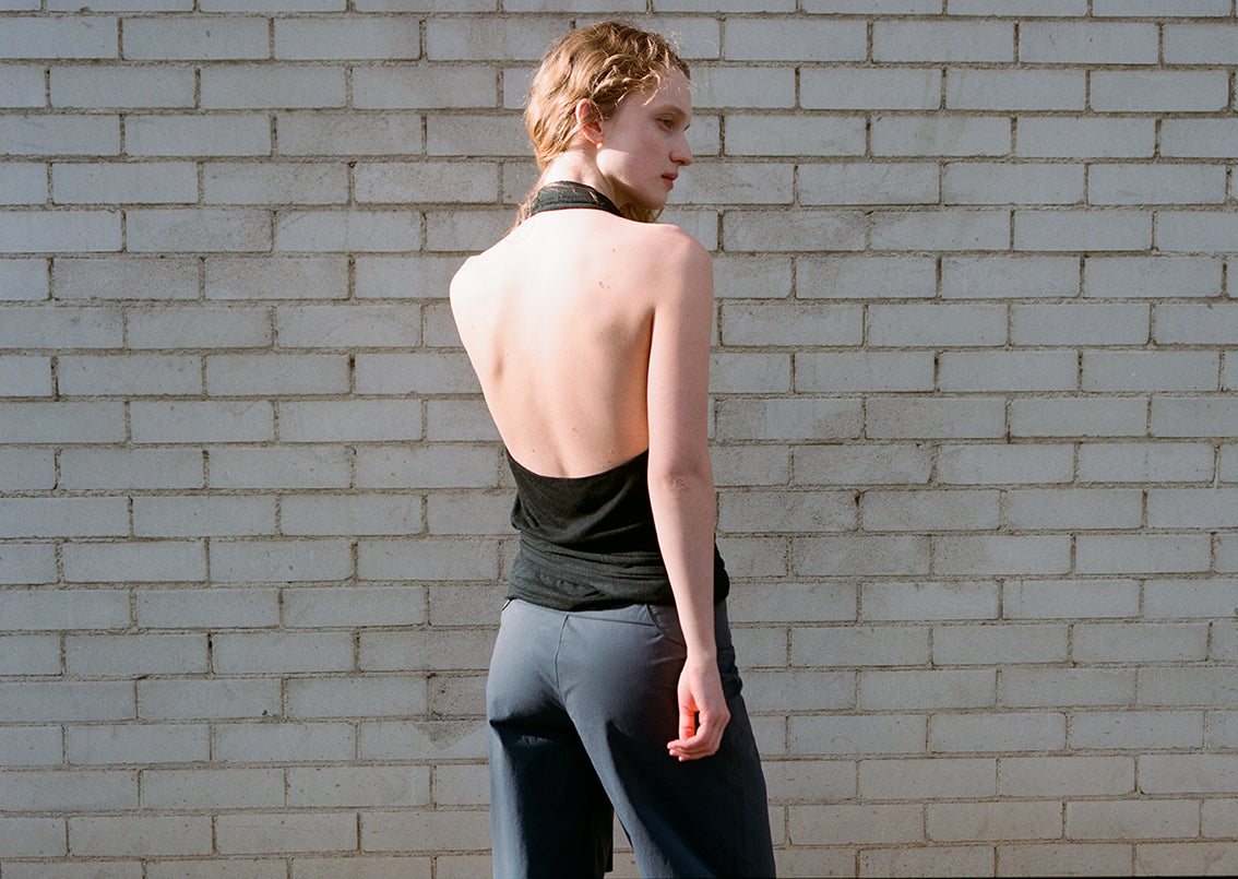 A person with short, light hair stands against a white brick wall, wearing a black backless top and the Panelled Pant - Charcoal by Deiji Studios in a straight-leg silhouette, their head turned to the side as they look over their shoulder.