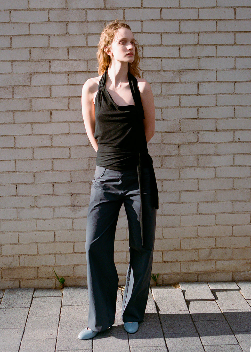 A woman with wavy hair stands against a light brick wall, wearing a black halter top and Deiji Studios Panelled Pant - Charcoal in a straight-leg silhouette. She completes the look with light blue flat shoes, gazing to the side, hands behind her back.