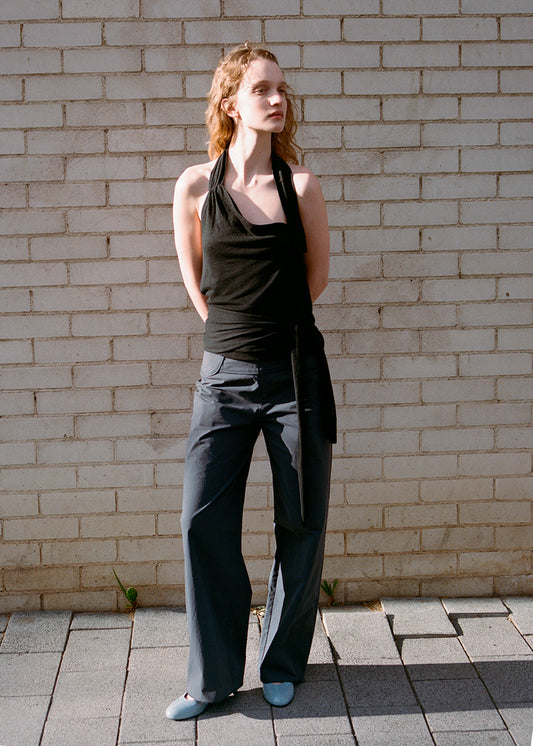A woman with wavy hair stands against a light brick wall, wearing a black halter top and Deiji Studios Panelled Pant - Charcoal in a straight-leg silhouette. She completes the look with light blue flat shoes, gazing to the side, hands behind her back.