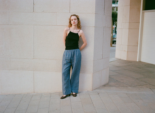 A person with curly hair, wearing a black tank top, Linen Ease Trouser - Azure Stripe by Deiji Studios, and black shoes stands against a light stone wall on a city sidewalk, looking at the camera with arms behind their back.